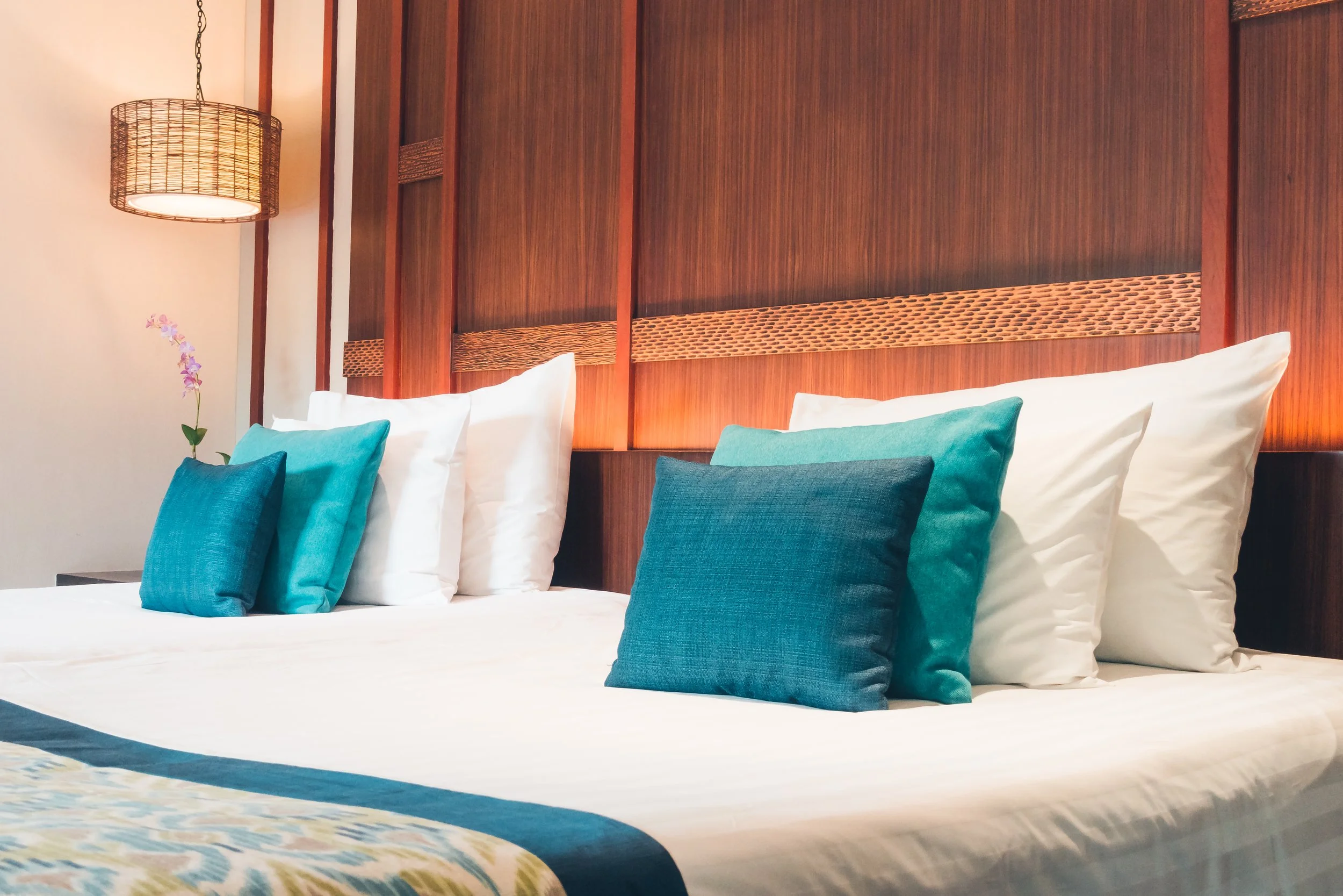 Un lit avec plusieurs coussins blancs et bleus dans une chambre avec panneau en bois derrière et une lampe suspendue.