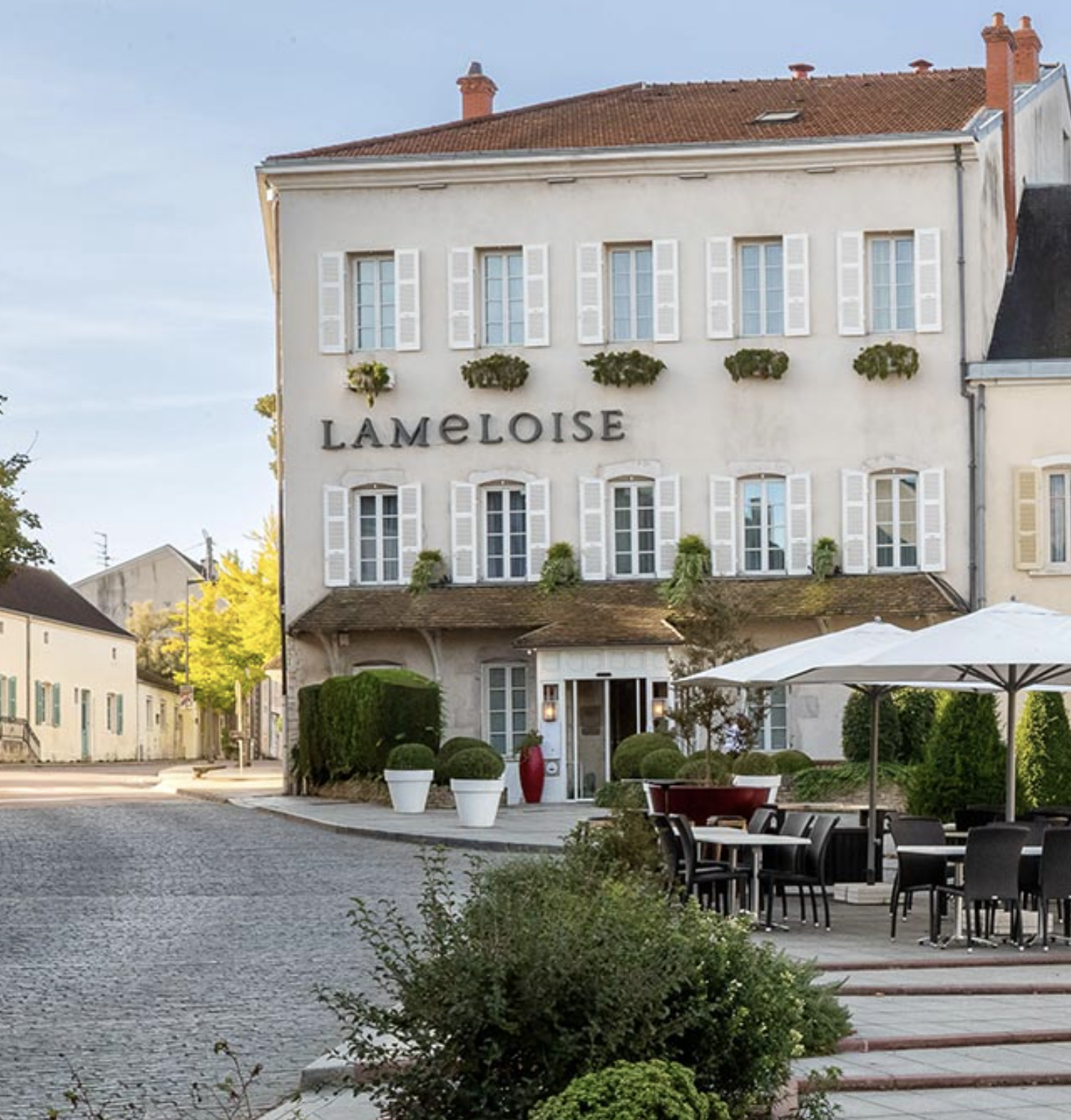 Un bâtiment blanc avec des fenêtres à volets blancs et le nom 'LAMELOISE' affiché sur la façade. Il y a une terrasse extérieure avec des tables et des parasols, le tout entouré de plantes et de buissons. Le ciel est clair, suggérant une journée ensoleillée.