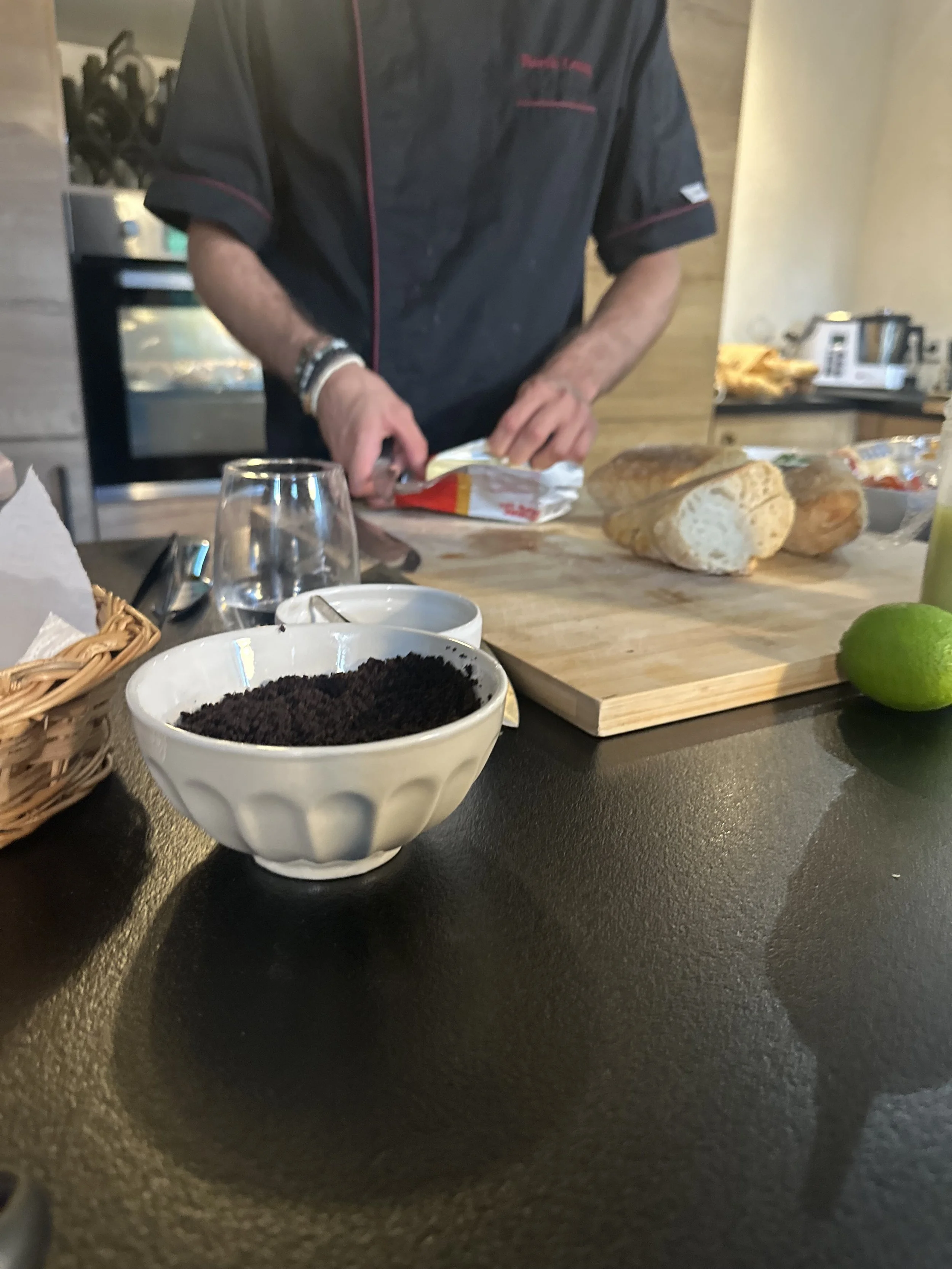Un chef prépare du pain sur une planche en bois, avec en premier plan un bol de café moulu, une lime, et un verre d'eau, le tout sur un comptoir noir.