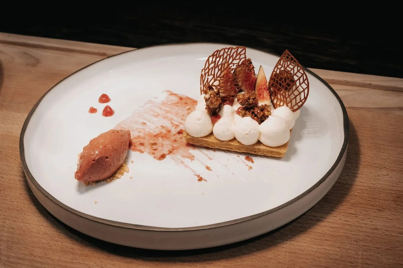 Gâteau de dessert avec des décorations en chocolat, figues, et crème, présenté sur une assiette blanche avec un sorbet à côté.