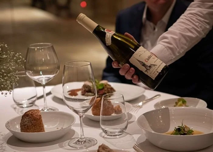 Une personne verse du champagne sur une table de restaurant avec plusieurs assiettes de nourriture, des verres à vin et de la vaisselle.