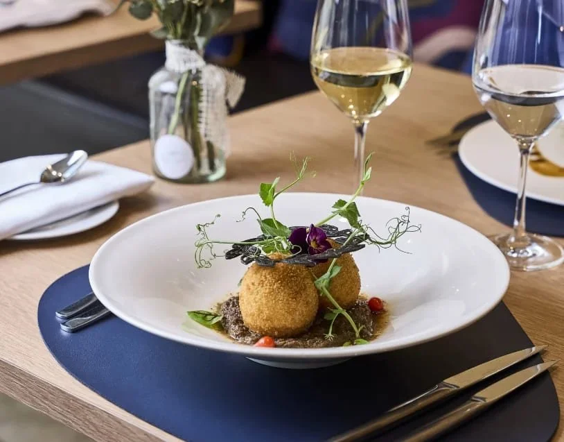 Un plat gastronomique avec deux boulettes frites, décoré de micro-pousses, garnitures et une sauce, accompagné de deux verres de vin blanc sur une table en bois.
