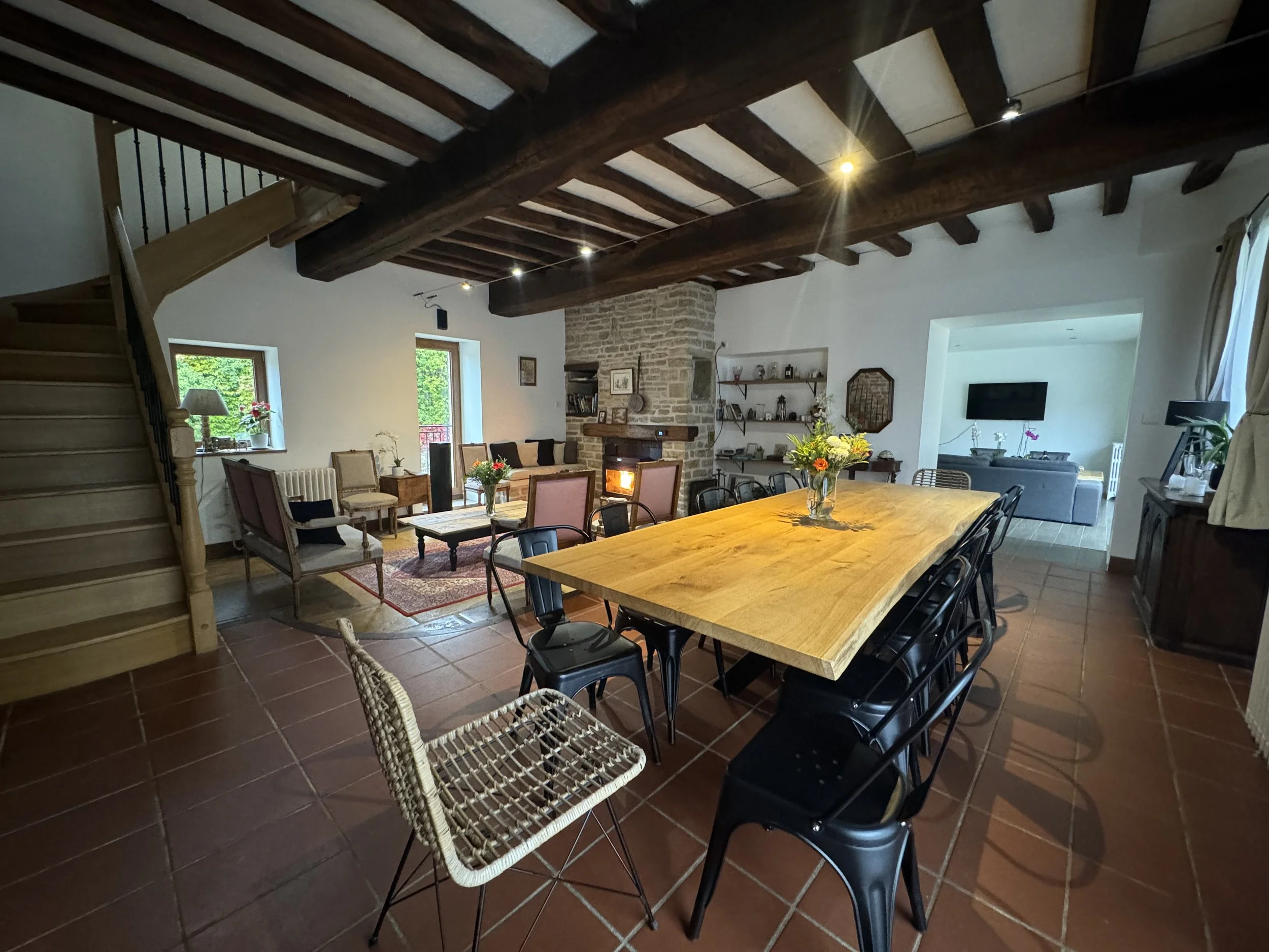 Une salle à manger spacieuse avec un grand coin salon. Au centre, une grande table en bois entourée de plusieurs chaises. À l'arrière, un coin cheminée avec quelques fauteuils et des étagères. La pièce a un plafond en poutres en bois et des fenêtres