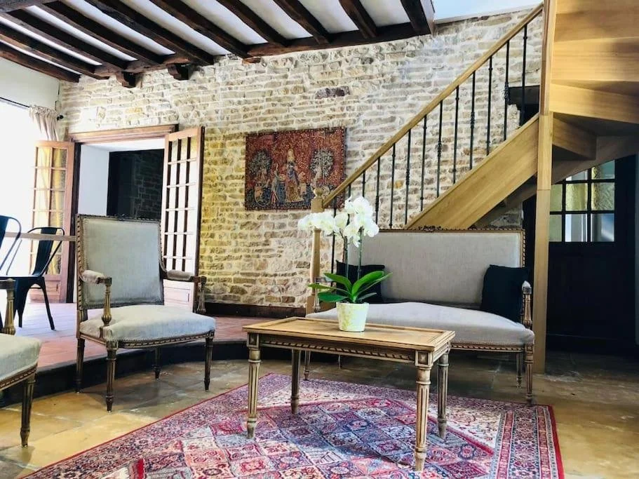 Salon intérieur avec un mur en pierre, un escalier en bois, un tapis oriental, une table en bois avec une plante, canapés en tissu clair et chaises.