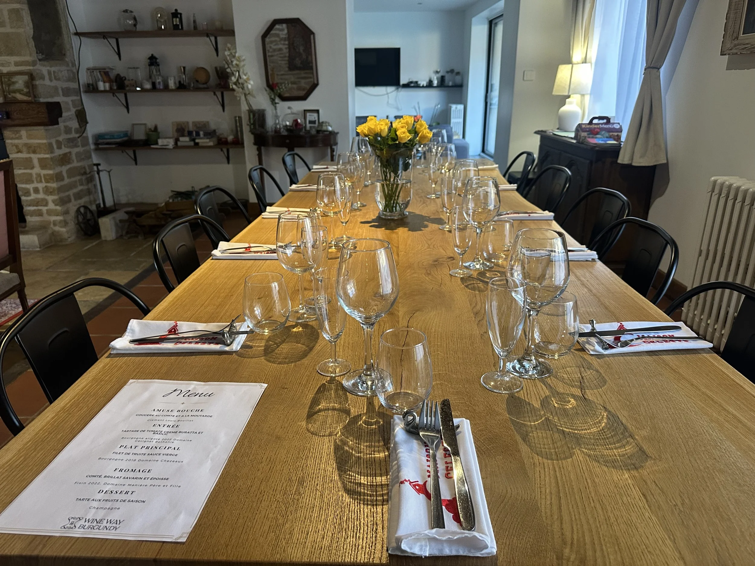 Table de dîner dressée avec plusieurs verres à vin, assiettes, couverts et une nappe blanche, au centre une vase avec des roses jaunes, dans un intérieur de maison avec une bibliothèque, un téléviseur et une cheminée.