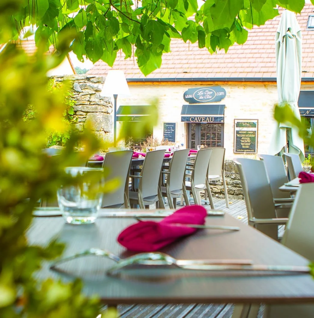 Une terrasse de restaurant en plein air avec des tables et des chaises, décorée avec des nappes roses, un verre d'eau, une fourchette, un couteau et un napperon rose. En arrière-plan, un bâtiment en pierre avec des enseignes en français, sous un arboré feuillu.