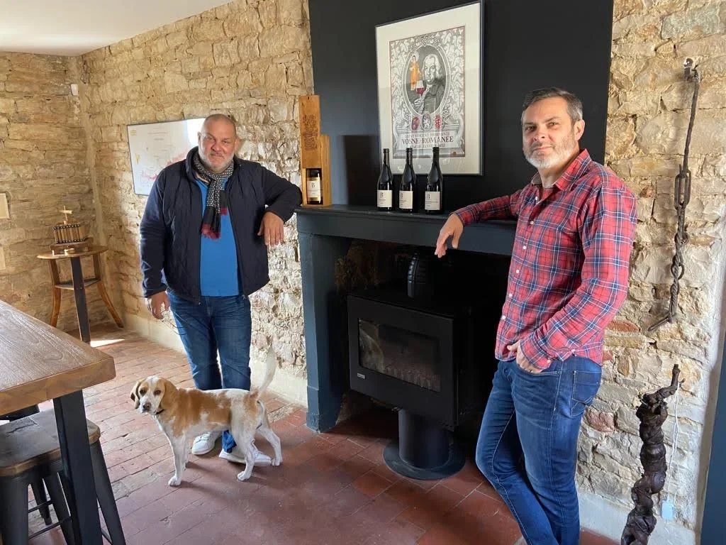 Deux hommes posent dans une pièce avec un mur en pierre. L'un d'eux est avec un chien blanc et marron à ses côtés, l'autre repose ses bras sur une cheminée décorée de bouteilles de vin et d'une affiche, dans une ambiance chaleureuse et décontractée.