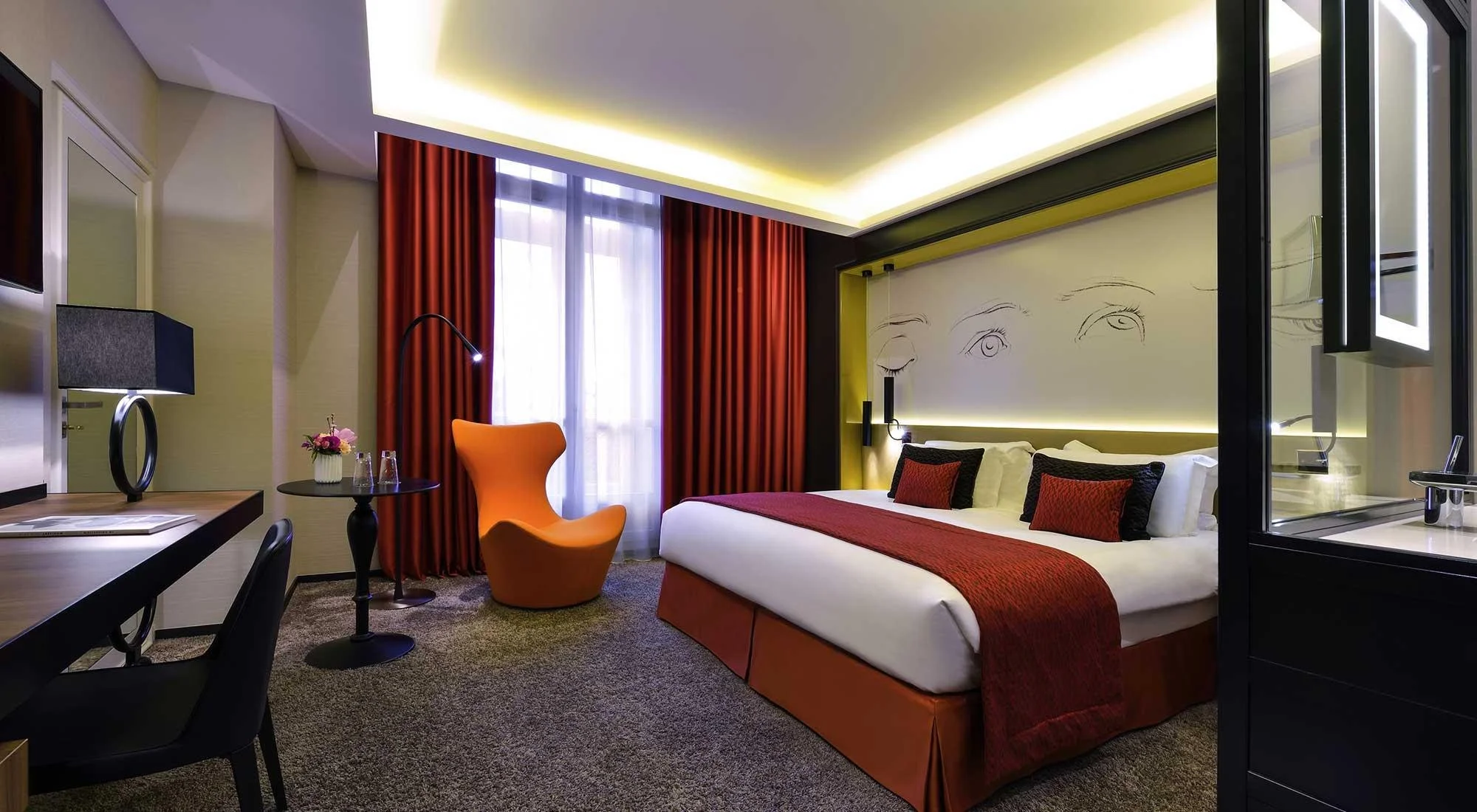 Chambre d'hôtel moderne avec lit, fauteuil orange, lampes, rideaux rouges et dessin minimaliste de visages sur le mur.