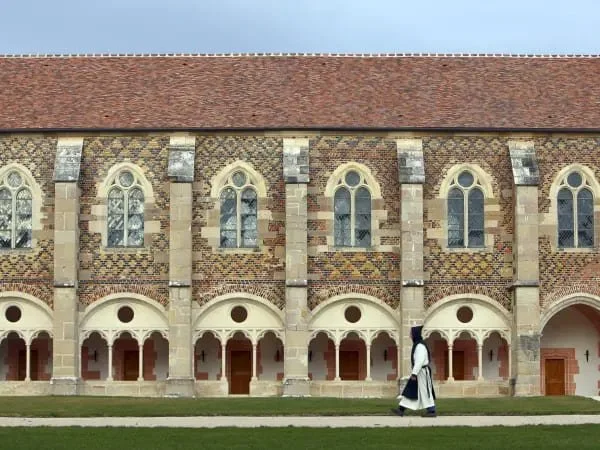 Une personne en habits traditionnels marche devant un bâtiment historique avec de grandes fenêtres en arc et une toiture en tuiles rouges.