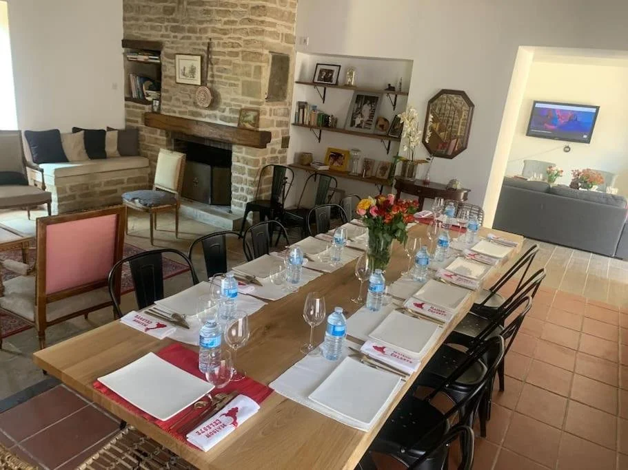 Salle à manger avec une grande table en bois, décorée de bouquets de fleurs, bouteilles d'eau, verres à vin, assiettes et couverts, dans une pièce avec cheminée en pierre, étagères avec livres et décorations, et un salon avec canapé et télévision.