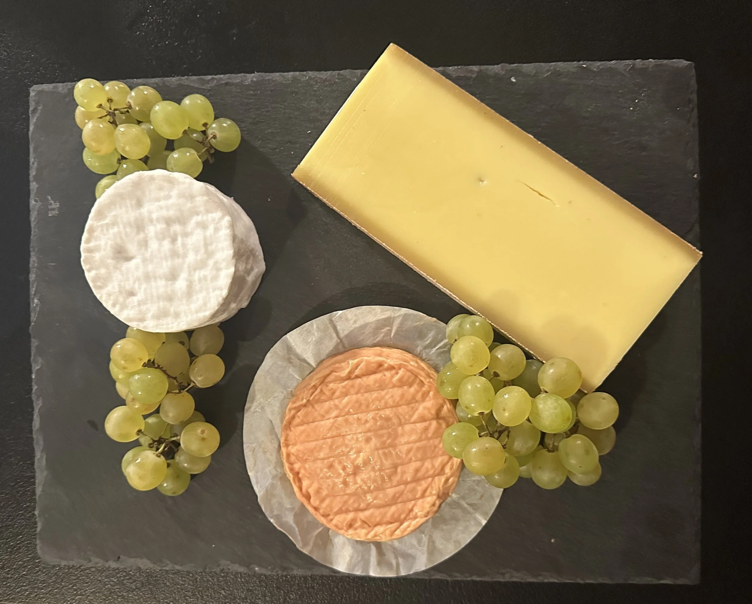 Plate fromages avec raisins blancs, fromage de chèvre, brie, et un morceau de fromage jaune dur.