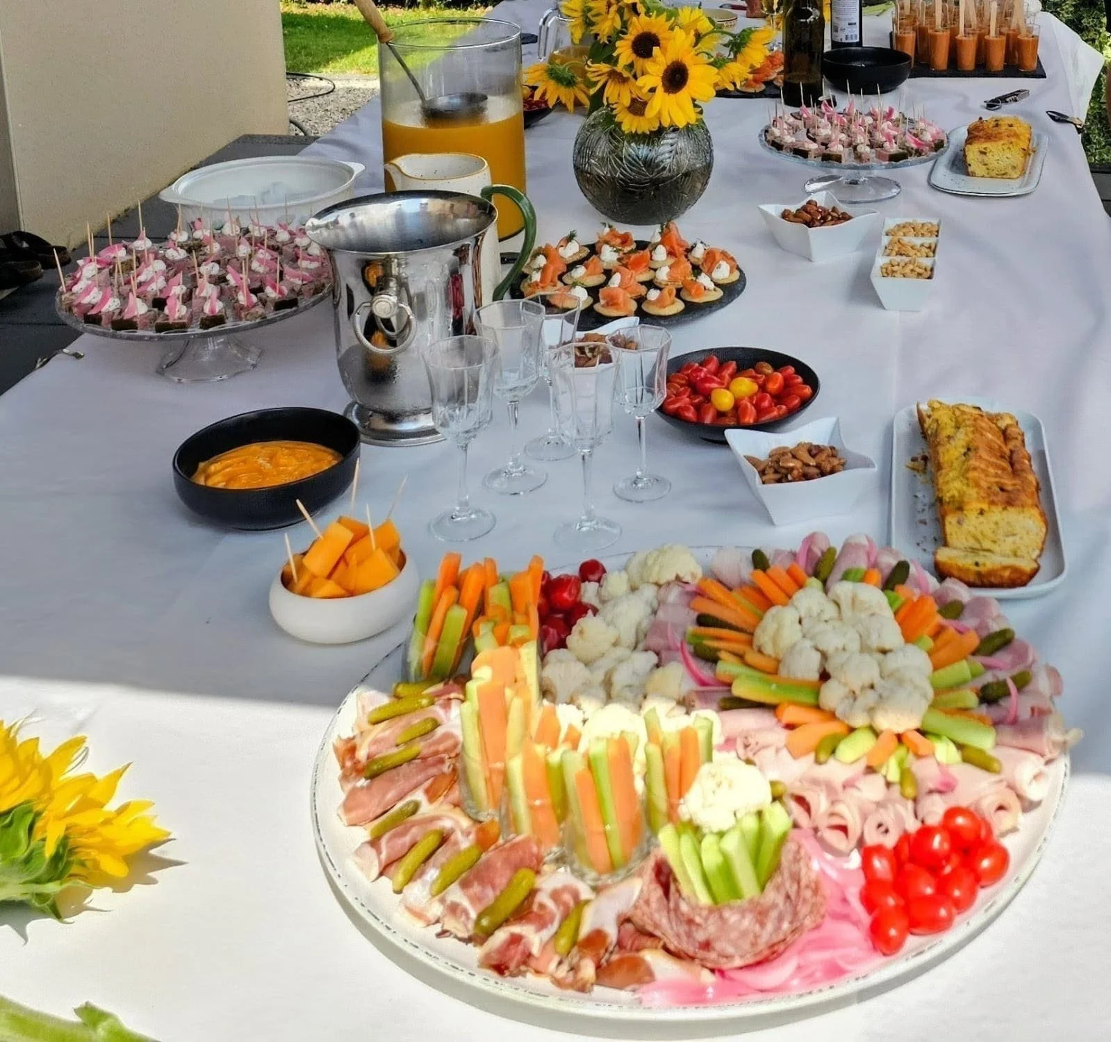 Table de fête avec assortiment de plats et amuse-bouches, comprenant un plateau de légumes décoratifs, des tomates, des snacks, un gâteau, et des boissons, décorée de fleurs jaunes.