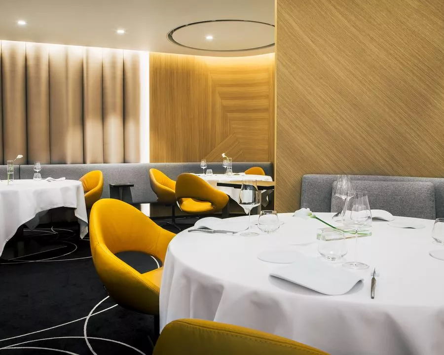 Salle à manger élégante avec tables rondes, nappes blanches, verres à vin, assiettes, couteaux, fourchettes, et chaises jaunes modernes dans un décor en bois et éclairage tamisé.