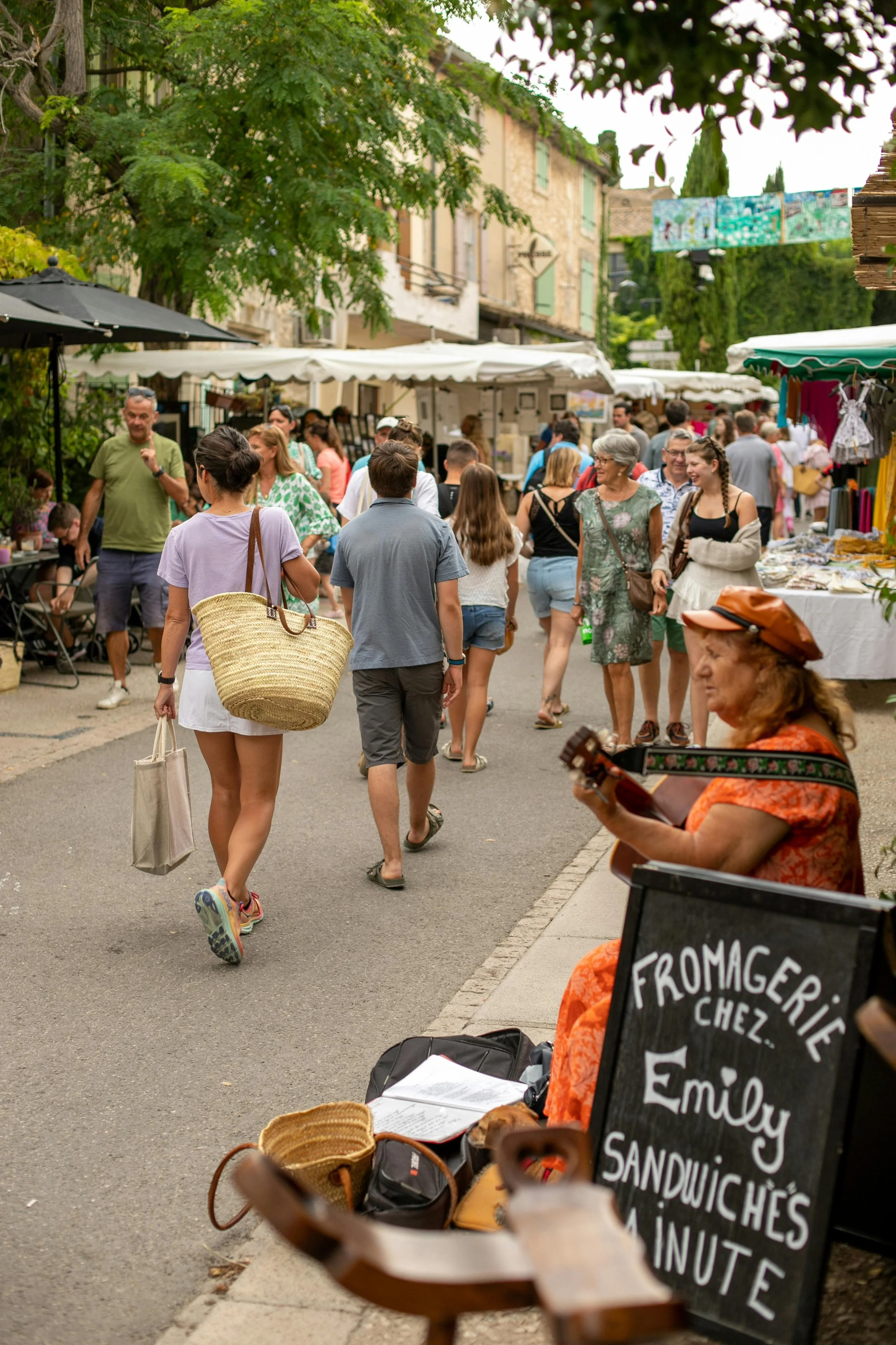 Scène de marché en plein air avec personnes marchant et stands de nourriture, musicienne jouant de la guitare, panneau indiquant 'Fromagerie Chez Emily'.