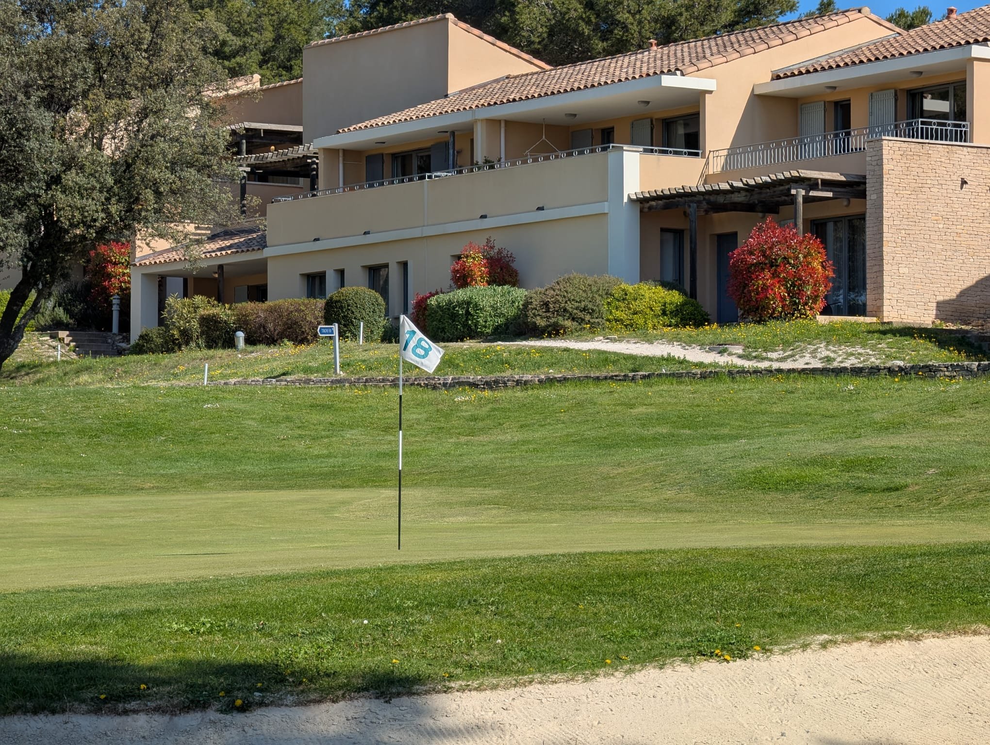 Terrain de golf avec drapeau indiquant le trou 18, pelouse verte et bâtiments résidentiels en arrière-plan, entourés de buissons et d'arbres.