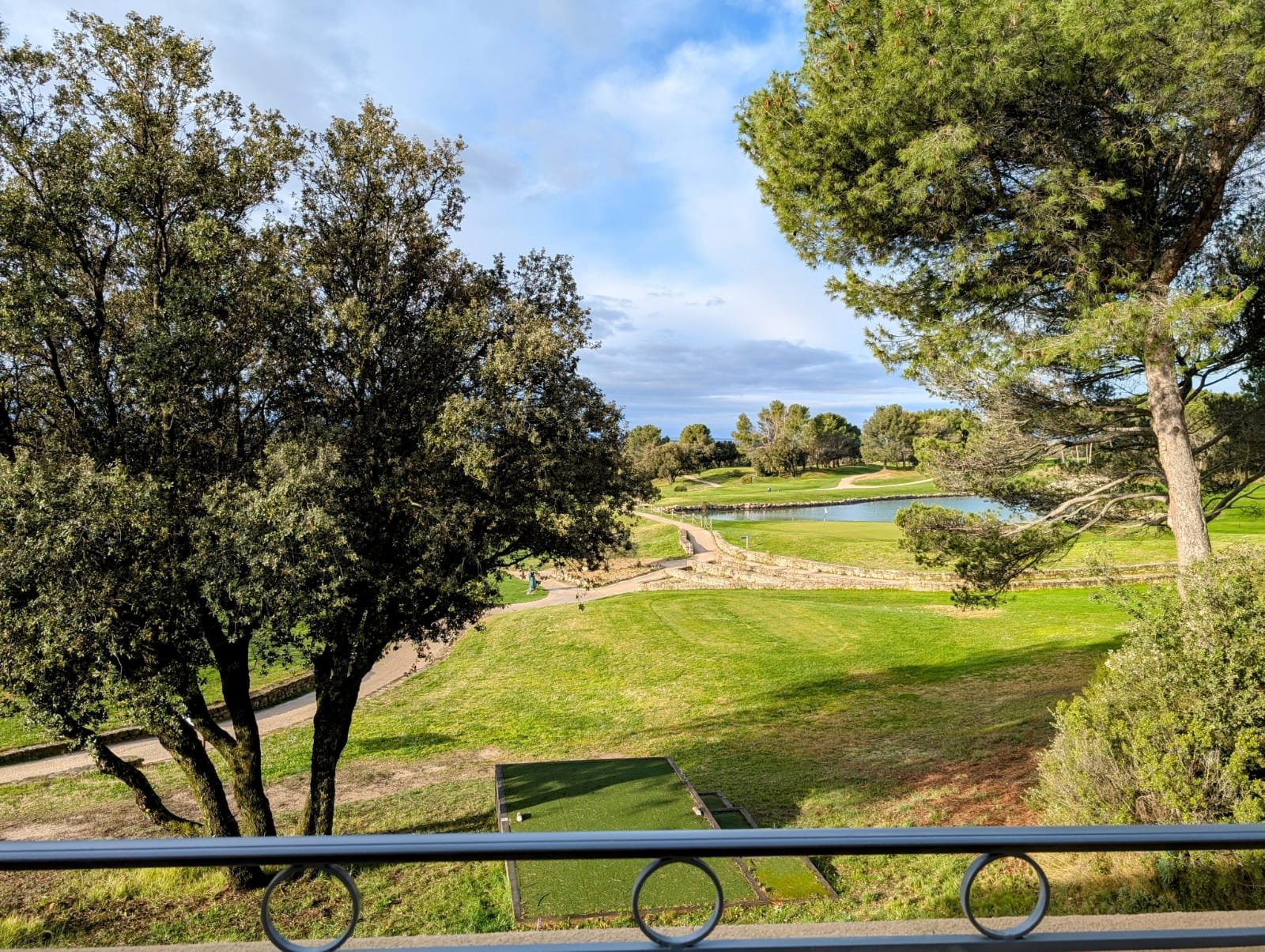 Paysage de terrain de golf avec arbres, pelouse verte et un petit étang sous un ciel partiellement nuageux.