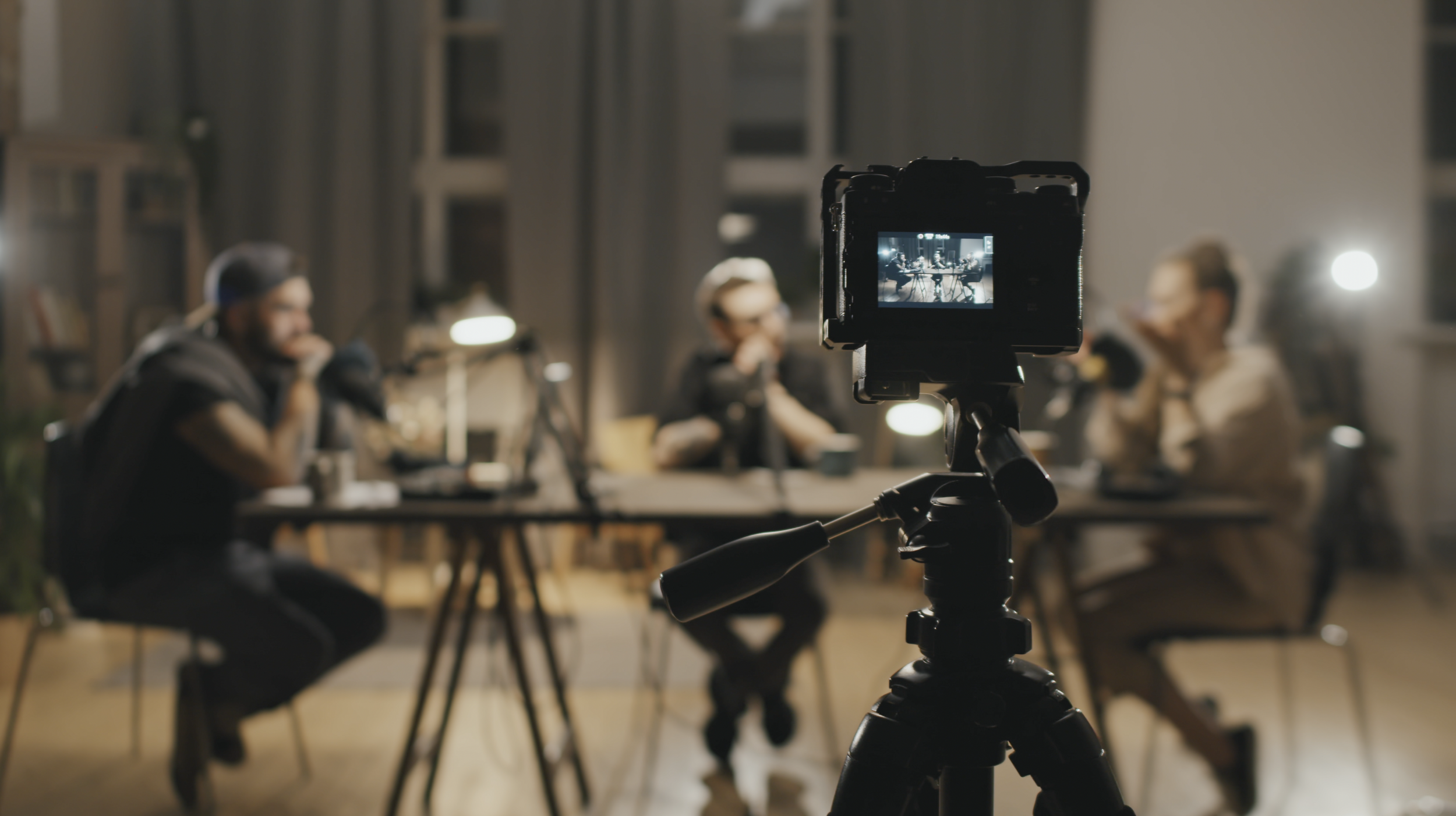 A podcast recording session with three people sitting at a table, speaking into microphones. A camera on a tripod is in the foreground, capturing the scene. The room is dimly lit with lamps, creating a cozy ambiance.