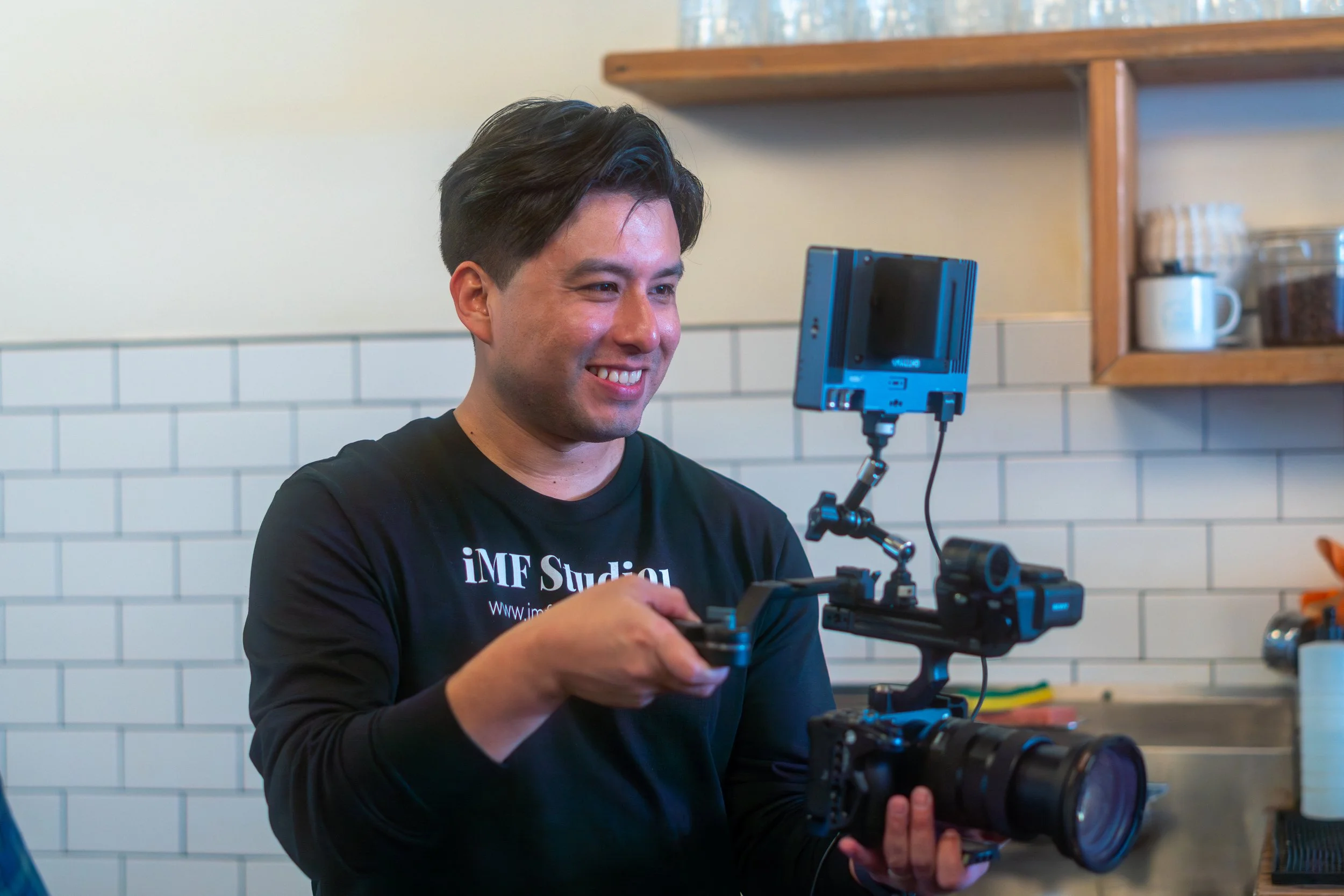 A young man with dark hair smiling as he looks at a professional camera setup with a mounted monitor in a kitchen with white tile backsplash and wooden shelves.