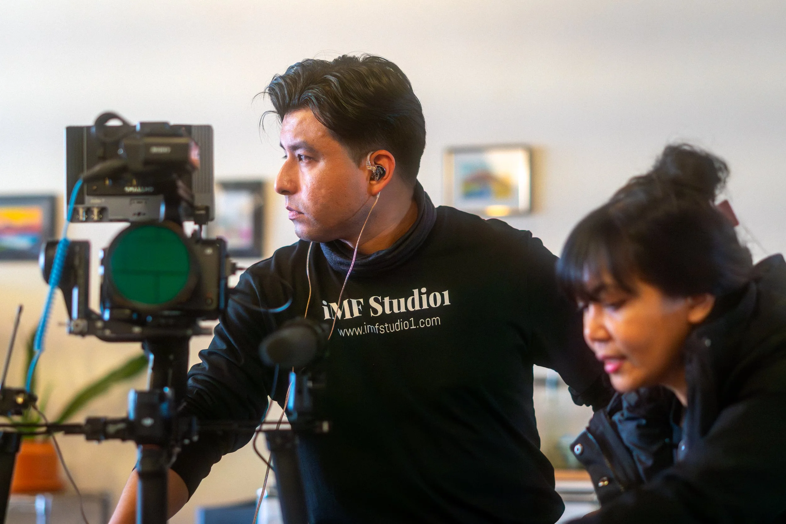Two people working on filming or photography, with one man looking intently at a camera mounted on a tripod and a woman beside him looking down, possibly adjusting equipment. They are indoors with framed pictures on the wall in the background.