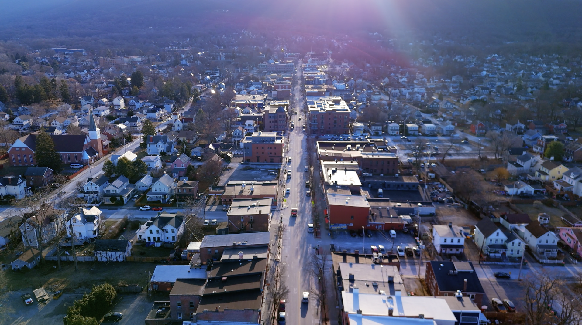 Aerial view of a small town with a central road lined with buildings, surrounded by residential houses and trees. The sky is clear with sunlight casting a glow over the scene.