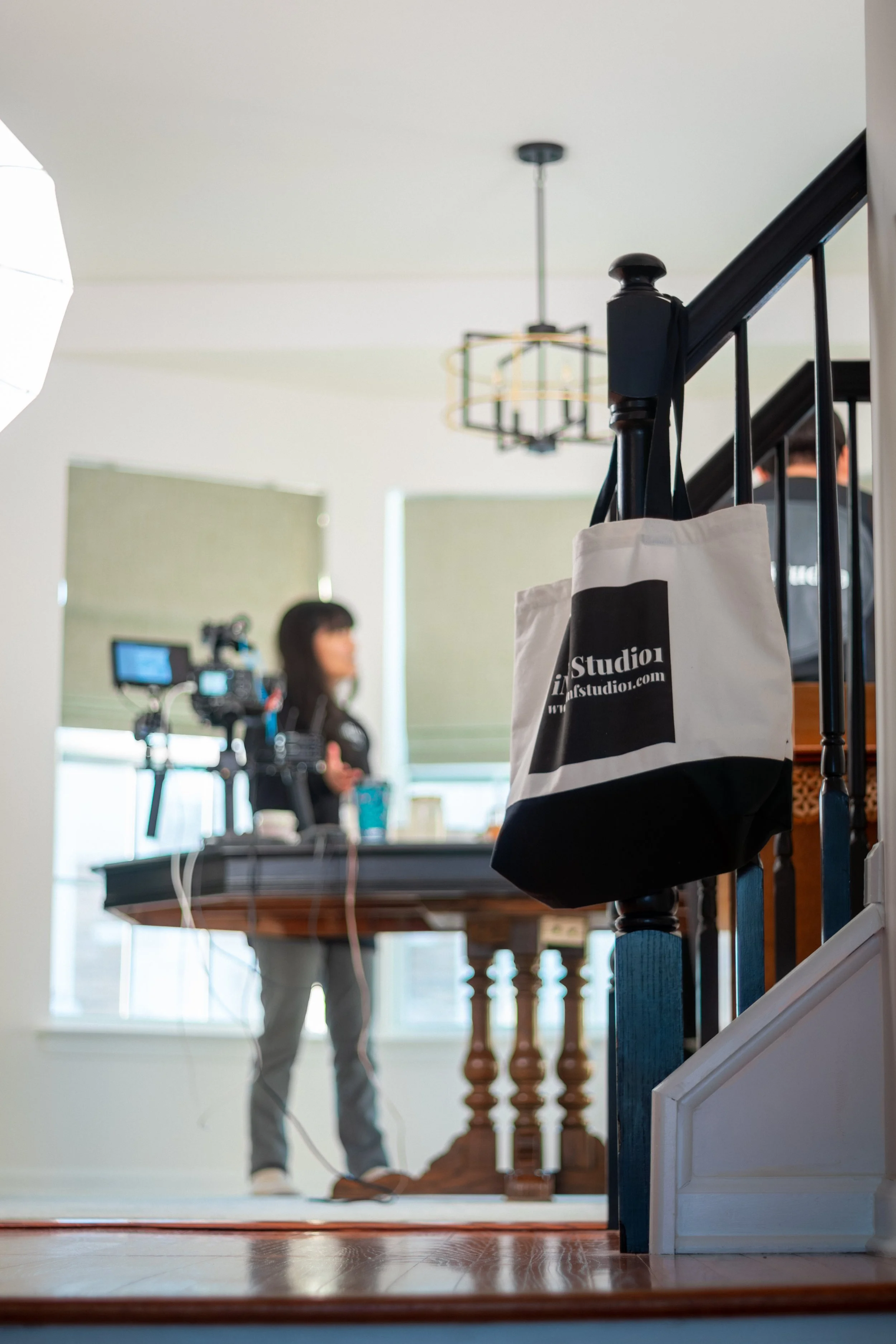 A woman with a camera and microphone in a room with large windows, a wooden table, and stairs with a black railing. A bag with the text 'Studio01' hangs on the railing.