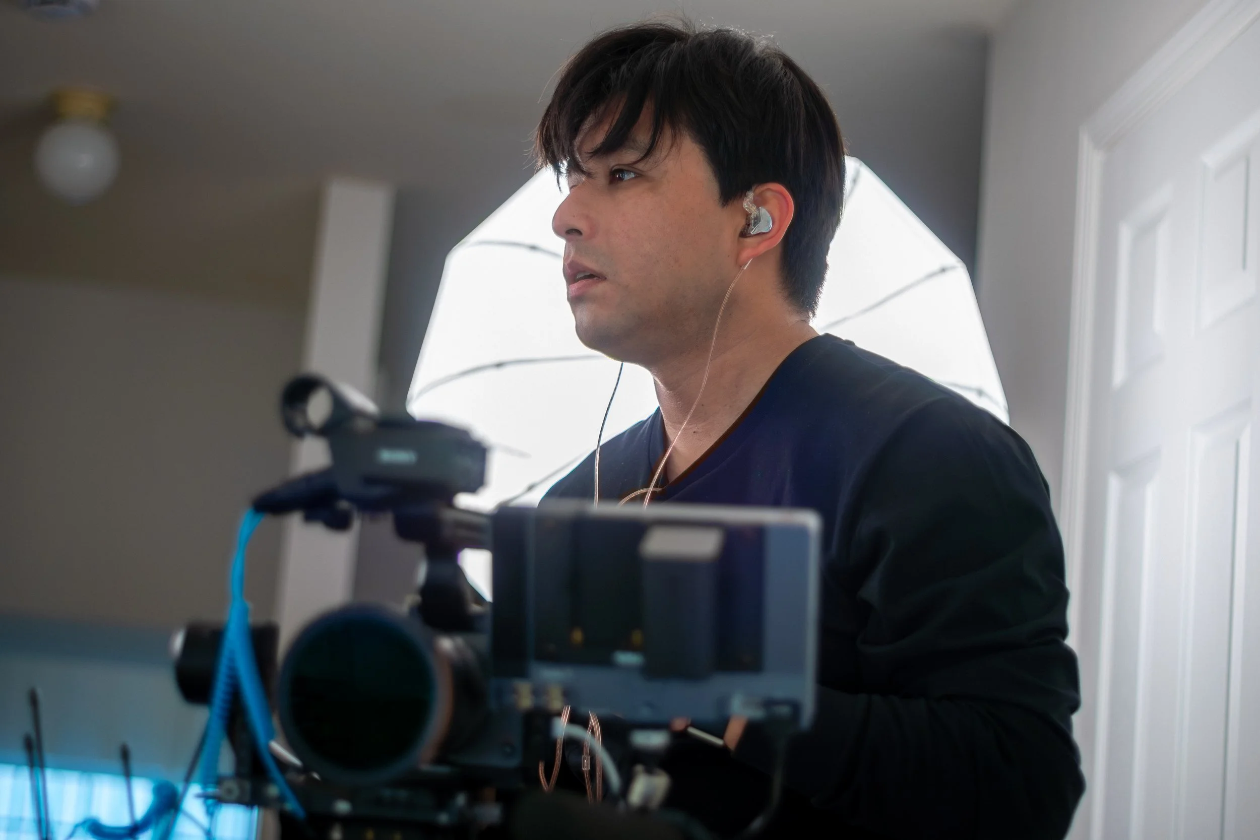 Man with dark hair wearing earbuds, looking to the side, in a room with a window and a door, working with a camera and equipment.