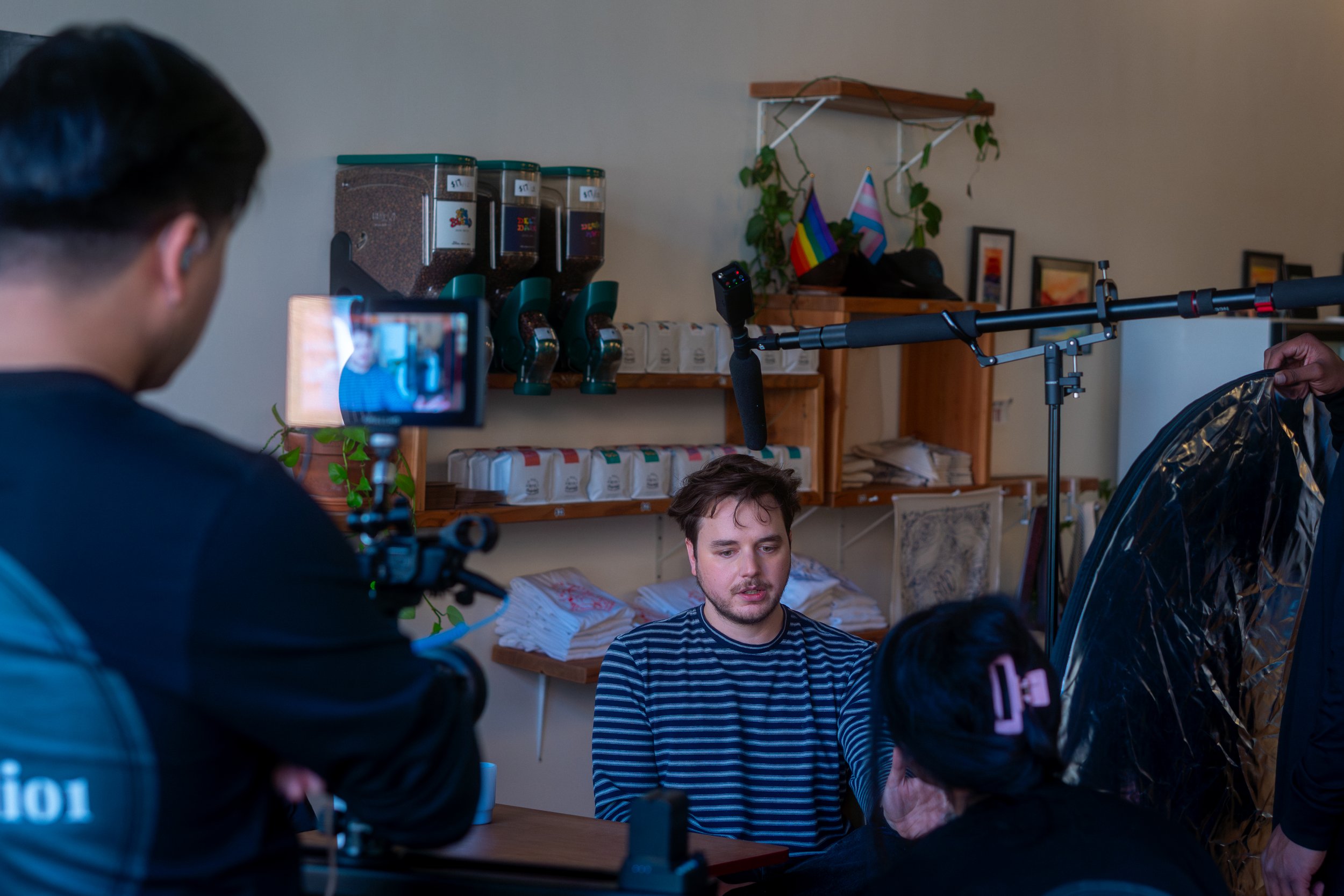 A man in a striped shirt is sitting at a table during a filmed interview, with a camera and crew members present, in what appears to be a cafe or small establishment.