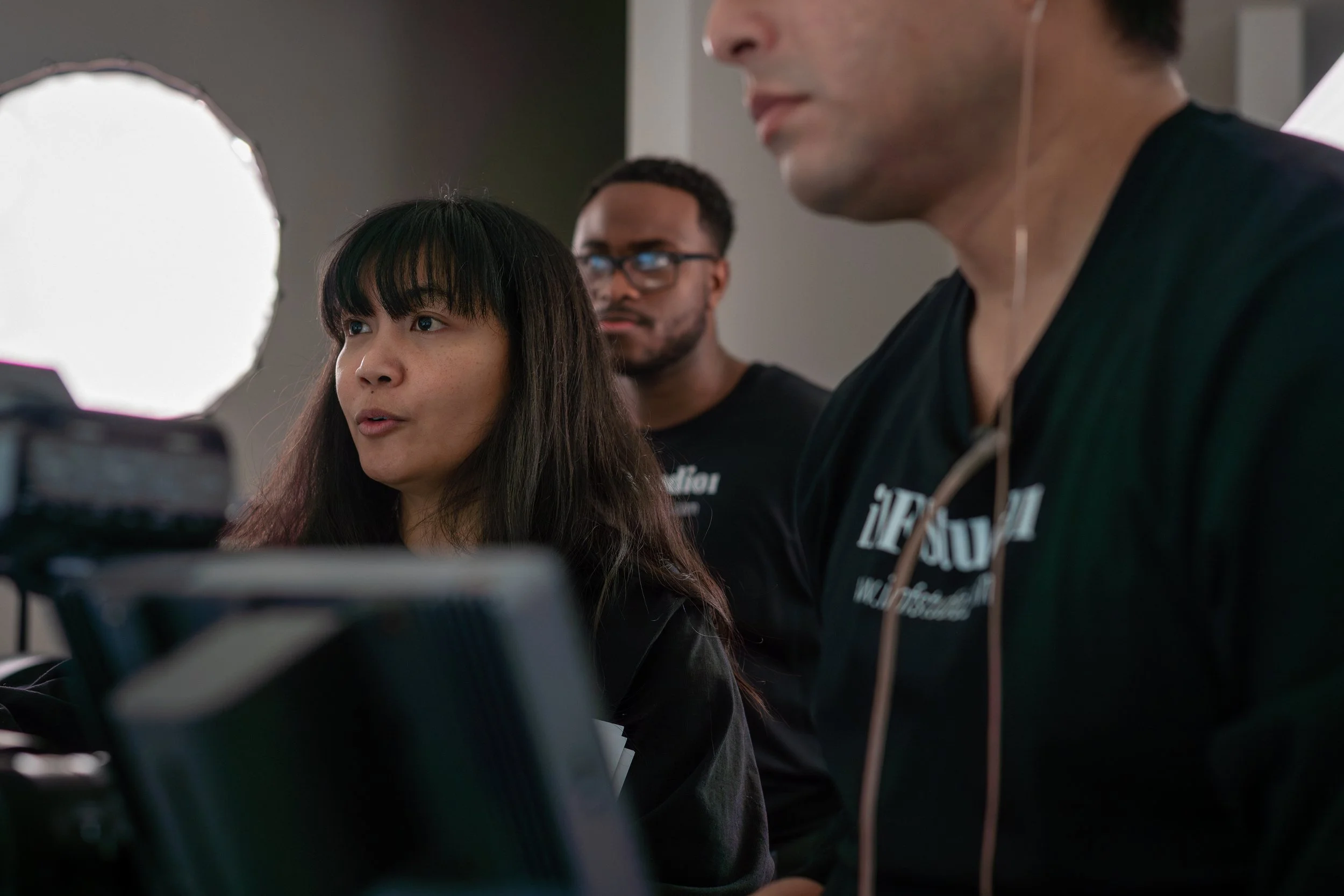 Three people working together in a technology setting, with a woman in front looking at a screen, and two men behind her, one with glasses and another wearing earbuds.