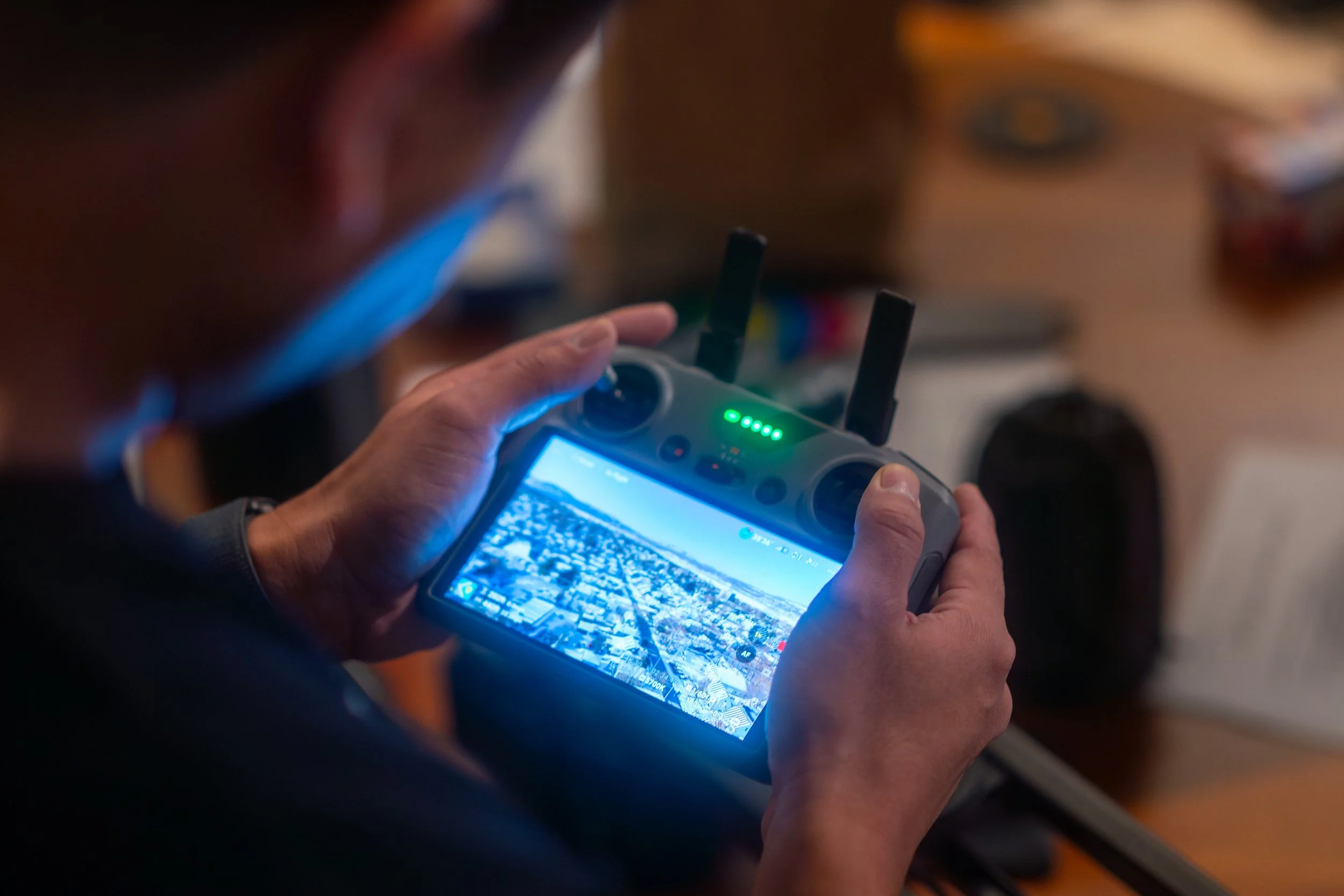 A person holding a drone controller with a live video feed of a city on the screen. The controller has two antennas and green LED indicators.