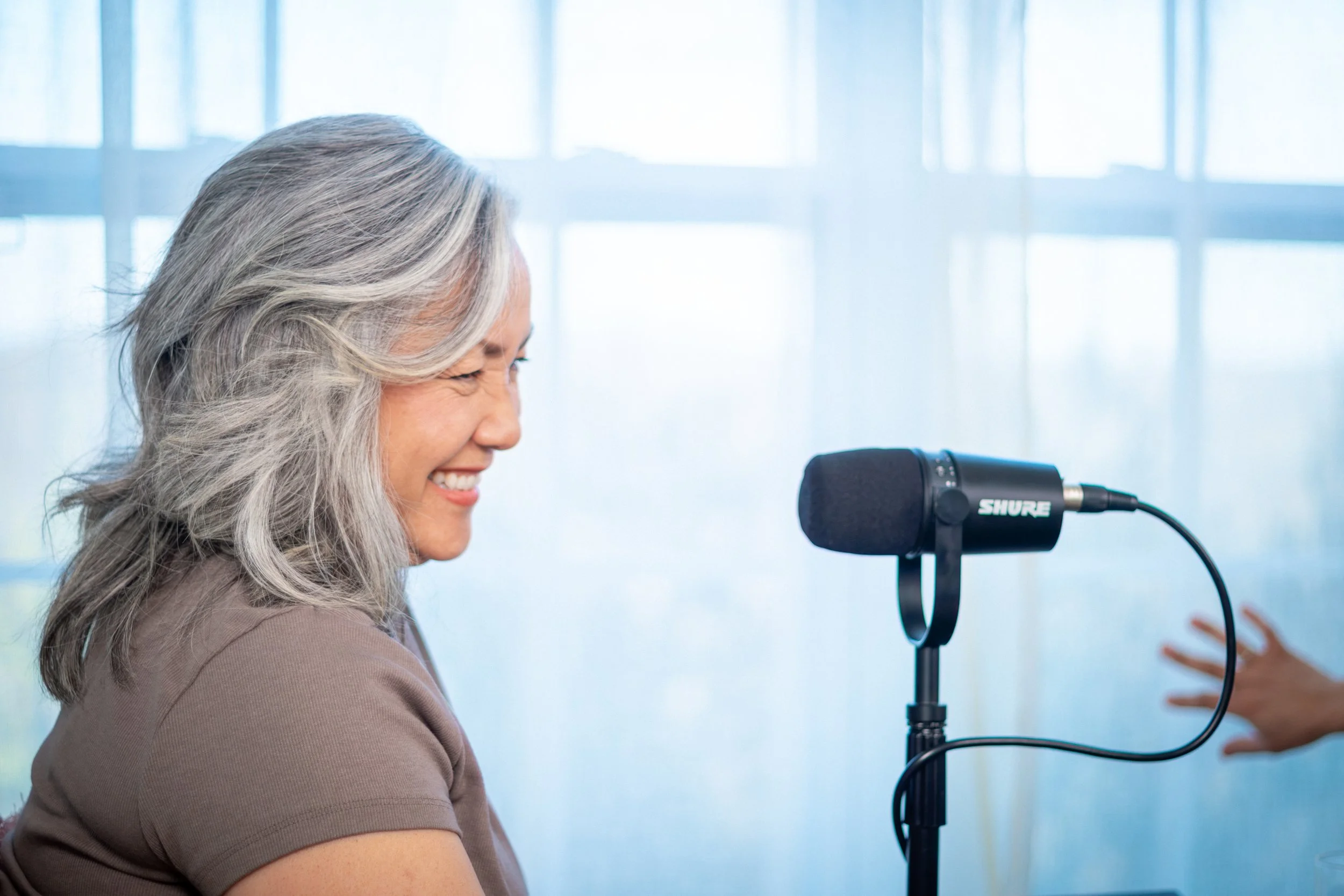A woman with gray hair smiling and talking into a Shure microphone during an interview or recording session.
