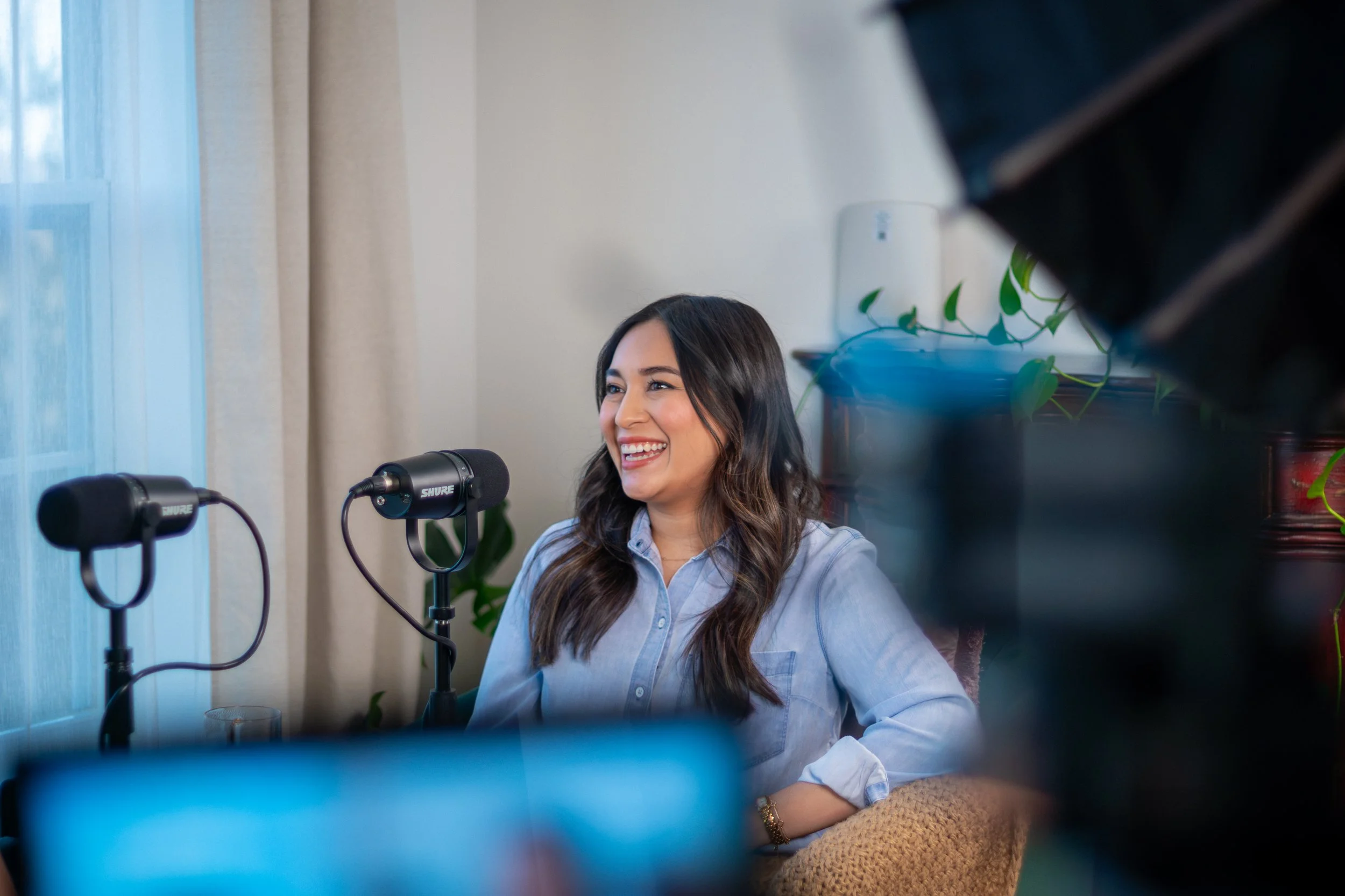 A woman with long dark hair in a light blue blouse sitting at a table, smiling and speaking into two microphones during a podcast recording, with plants and a window in the background.