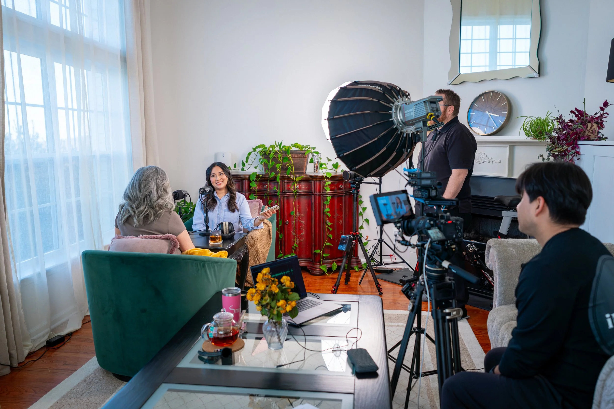 People filming an interview in a living room, with cameras, lights, and a crew present. Two women sit at a table with coffee, and one woman is speaking while the other listens.