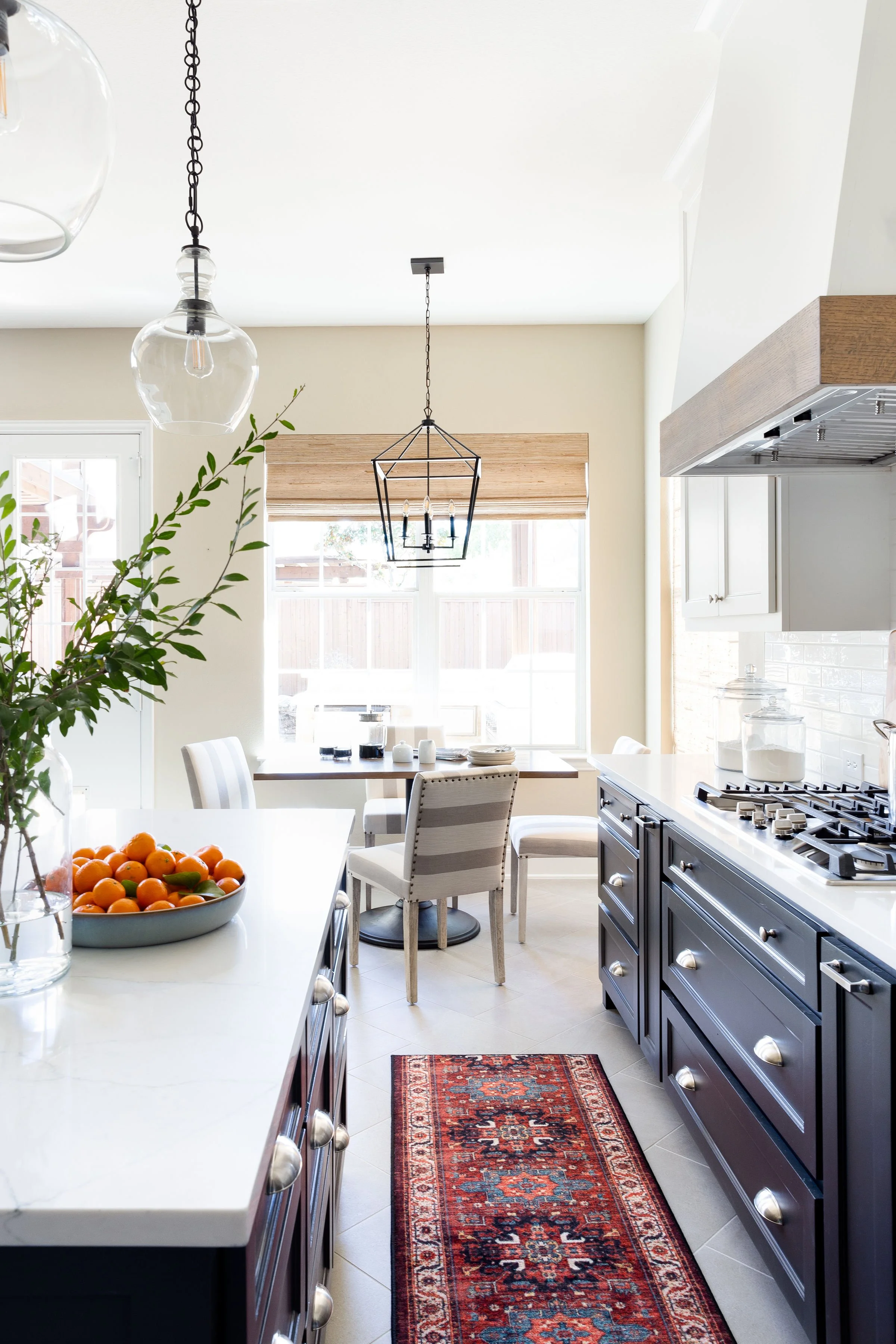 Modern kitchen with white countertops, dark cabinets, and a decorative rug. A bowl of oranges, green branches in a vase, and a dining area with chairs are visible. Pendant lighting hangs from the ceiling.