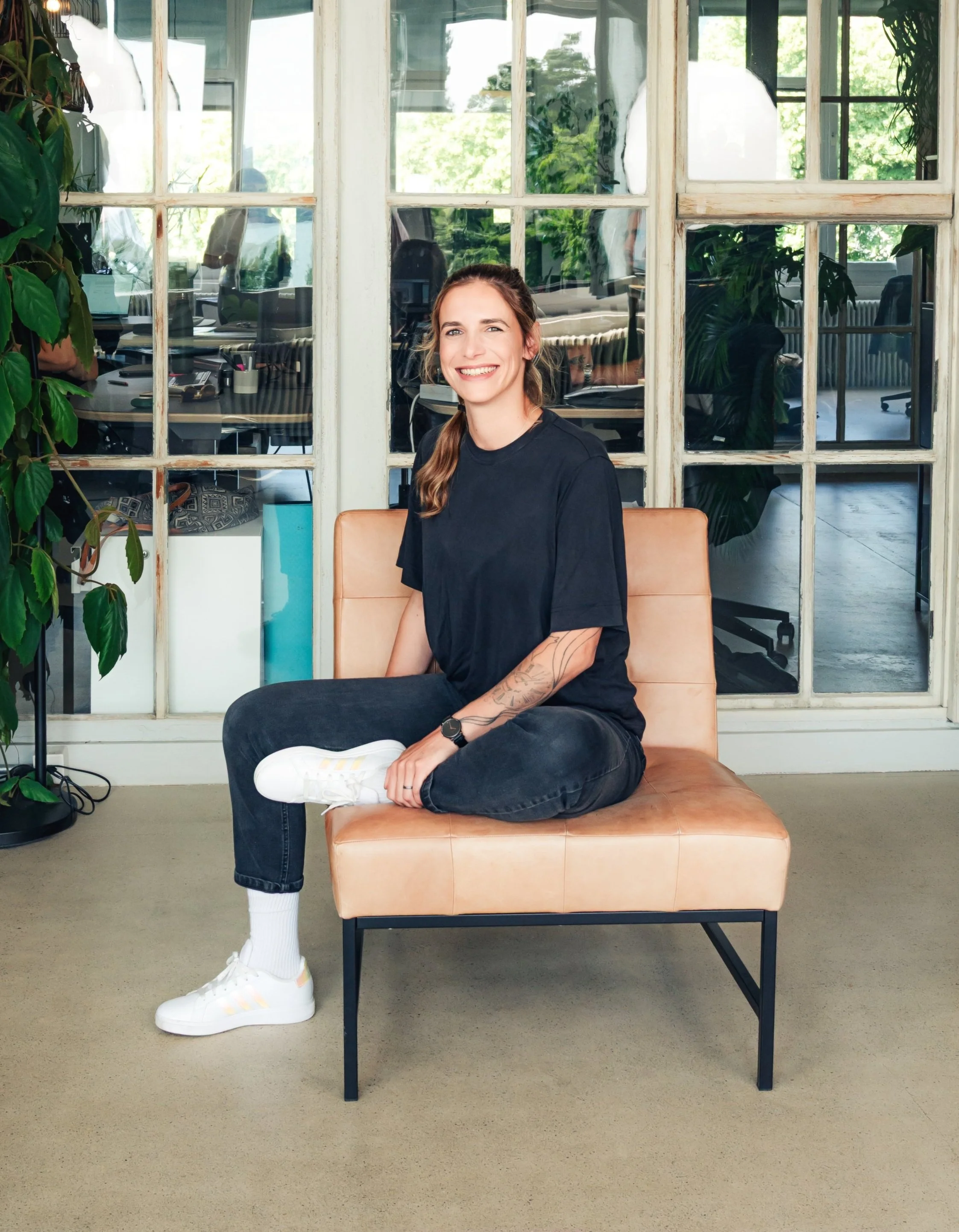 Junge Frau sitzt auf einer Couch in einem modernen Büro, trägt schwarze Kleidung und weiße Sportschuhe, lächelt und schaut fröhlich in die Kamera.