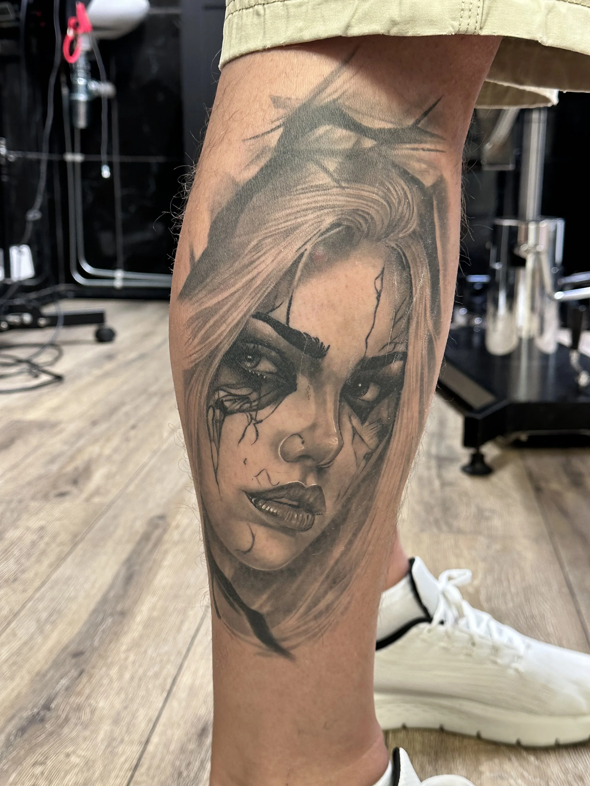 Tattoo of a woman’s face with detailed eyes and lips inked on a person's calf, with parts of hair and a branch or twig visible.