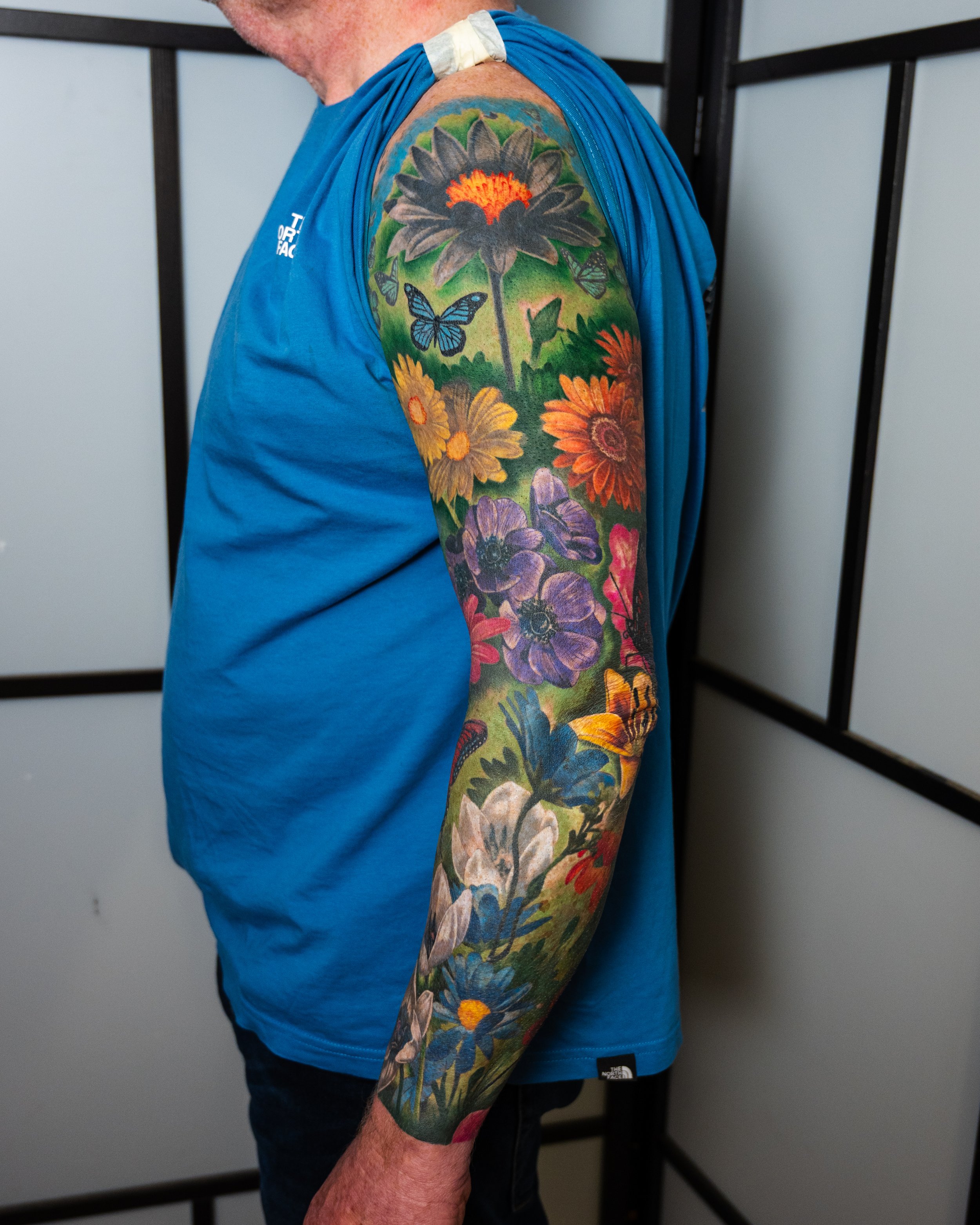 A man with a colorful floral tattoo sleeve on his left arm, featuring large flowers and butterflies, wearing a blue sleeveless shirt, standing indoors in front of a white wall with black framing.