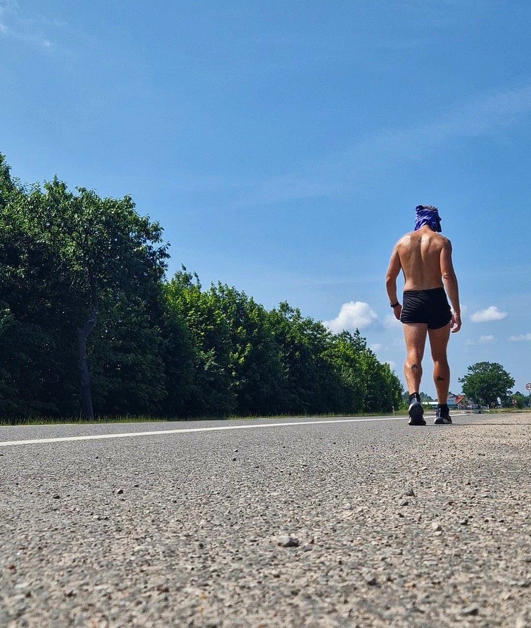 Back view of a shirtless person with a Super Mindset walking on a road, wearing shorts and shoes, surrounded by trees under a clear blue sky.