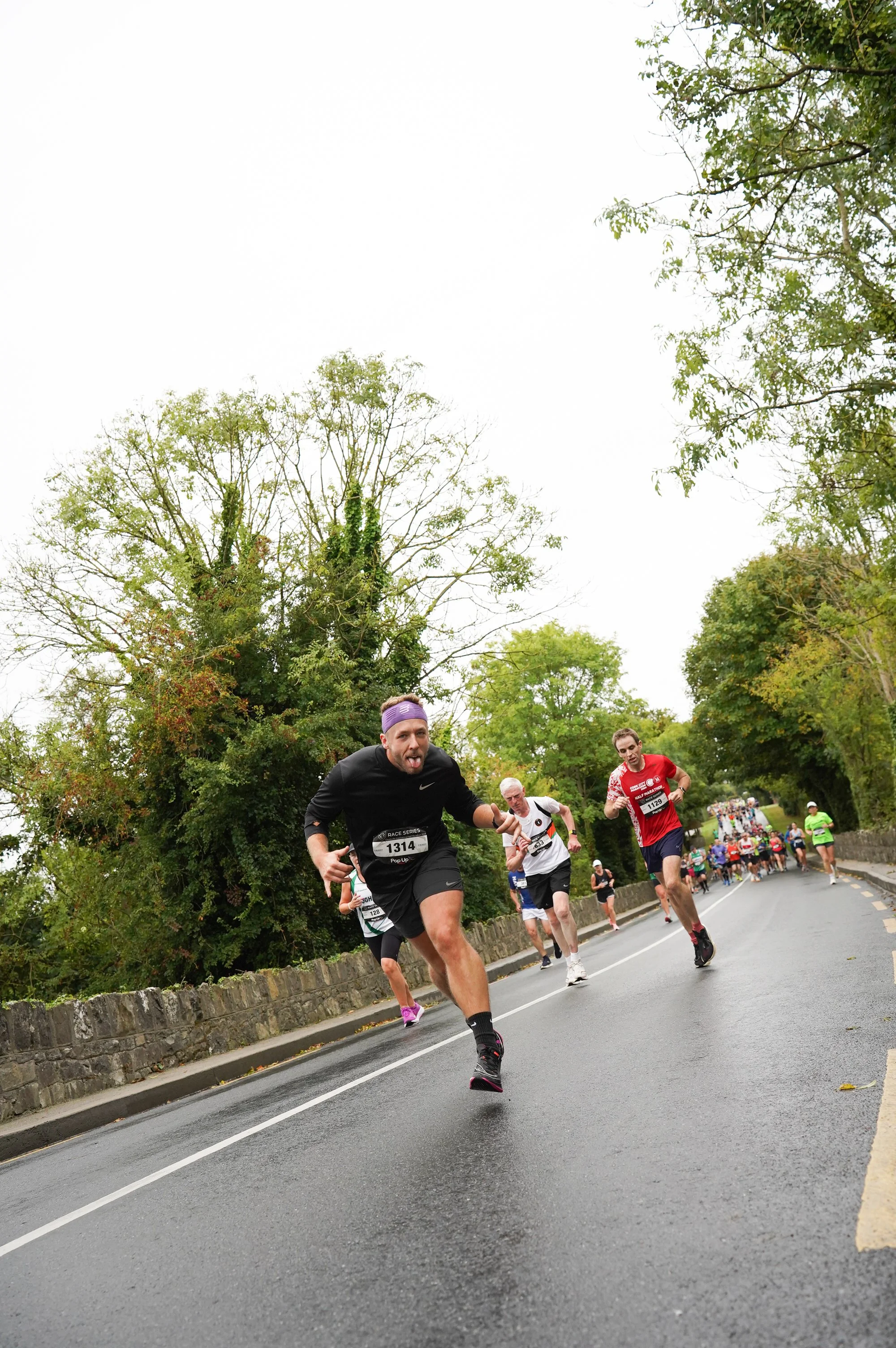 Runners participating in a road race on a tree-lined street, with a leading runner in black stretching out and others following closely, under overcast skies.
