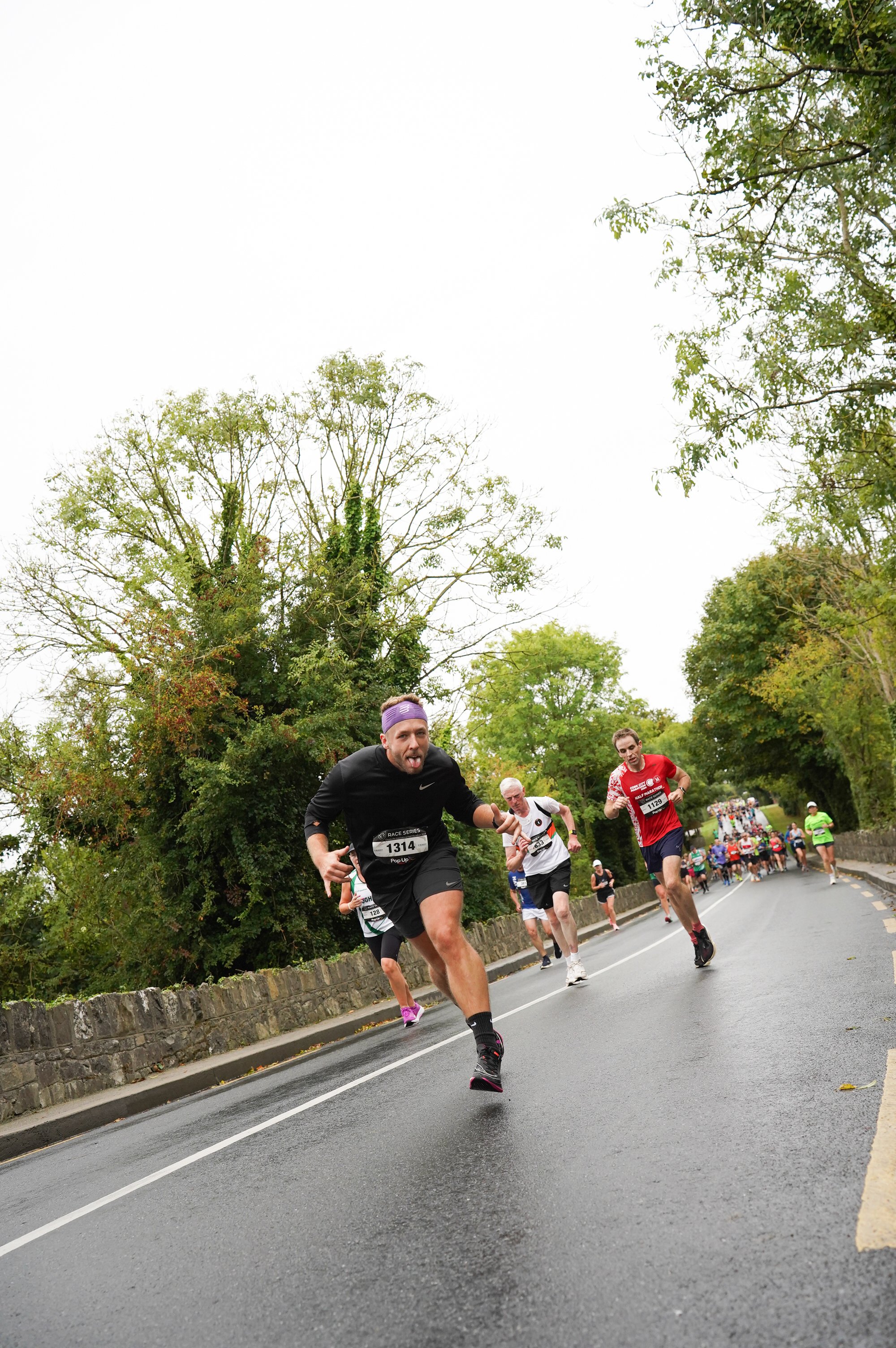 Runners participating in a road race on a tree-lined street, with a leading runner in black stretching out and others following closely, under overcast skies.