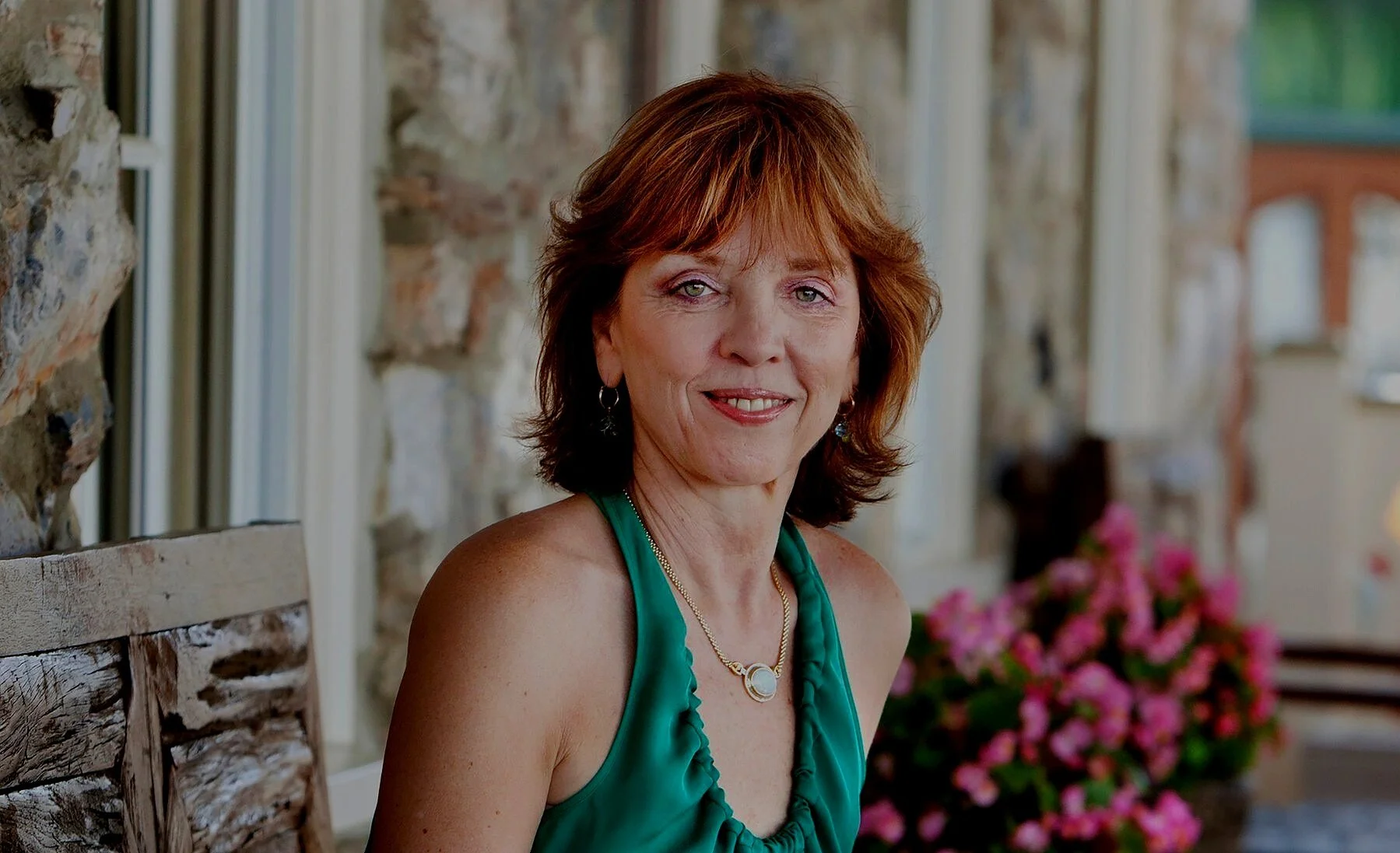 Writer Nora Roberts with auburn hair wearing a green sleeveless top and a necklace, sitting on a wooden chair with a stone wall and pink flowers in the background.