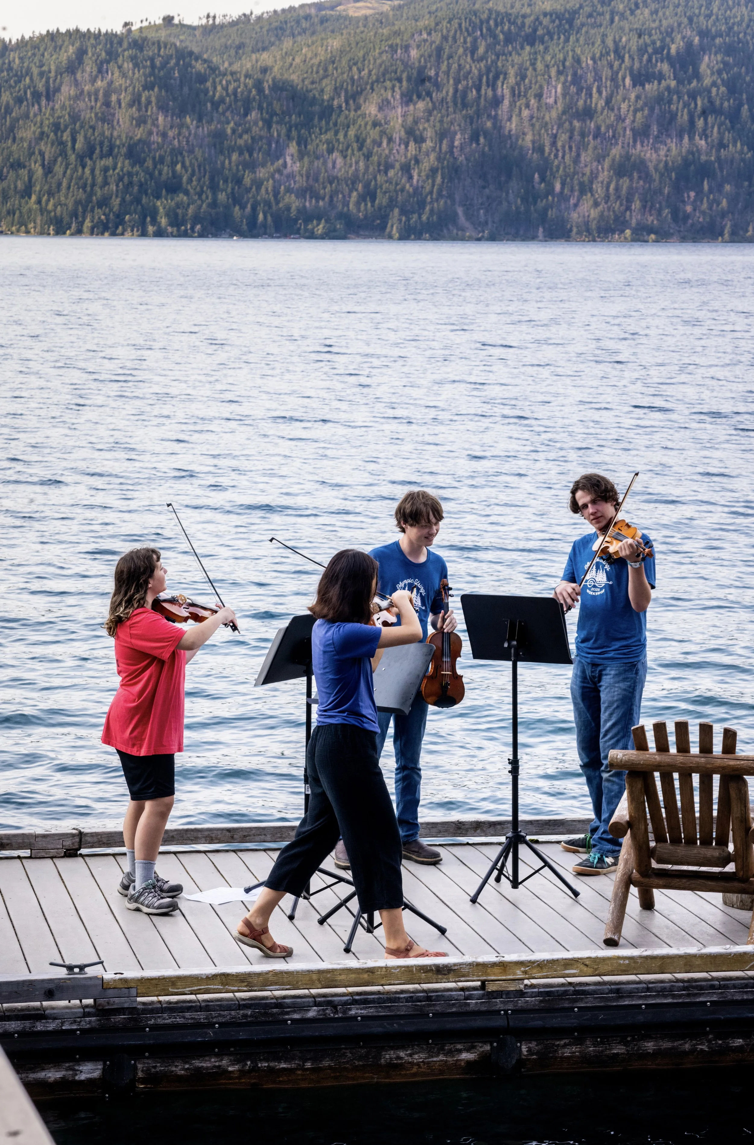 A group of five young musicians playing violins and a viola outdoors on a wooden dock next to a lake, with a forested mountain in the background.