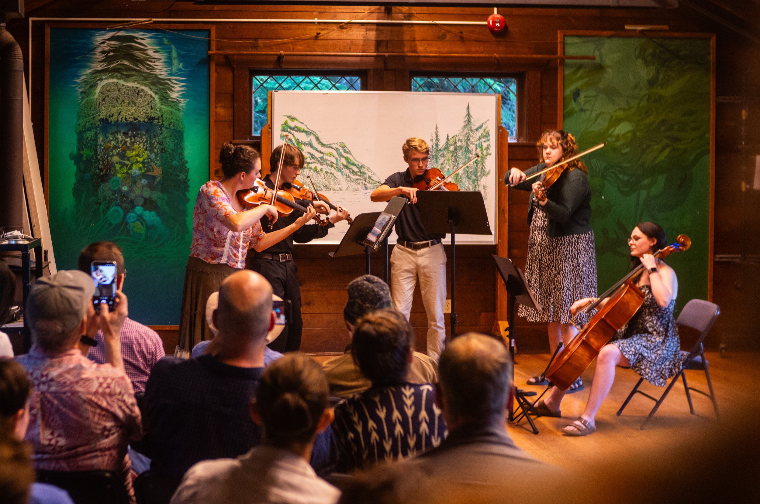 A group of five musicians performing in a rustic indoor setting with wooden walls, surrounded by colorful nature-themed artwork. Four musicians are playing violins, with a woman playing a cello, while a woman stands and plays the violin. The audience is seated and watching, some recording with phones.