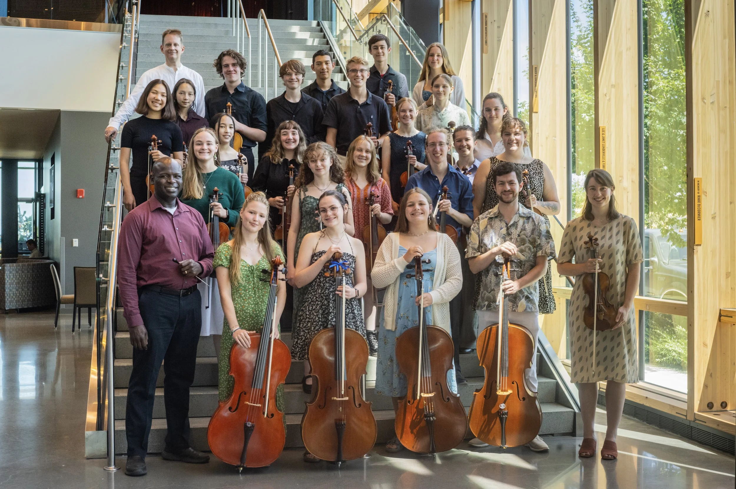 A group of young musicians holding cellos andviolin posing on stairs inside a modern building with large windows showing trees outside.