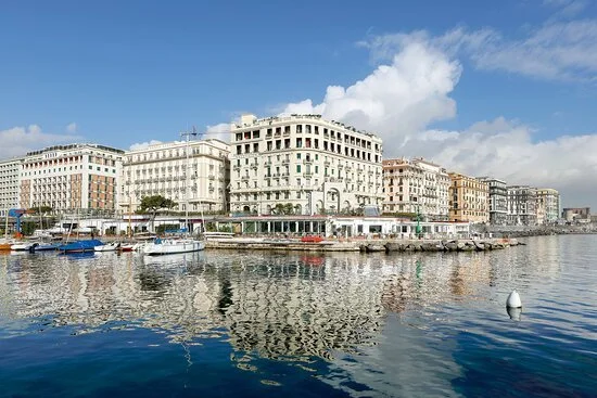 A waterfront cityscape with historic buildings, boats docked, and calm water reflecting the buildings under a partly cloudy sky.