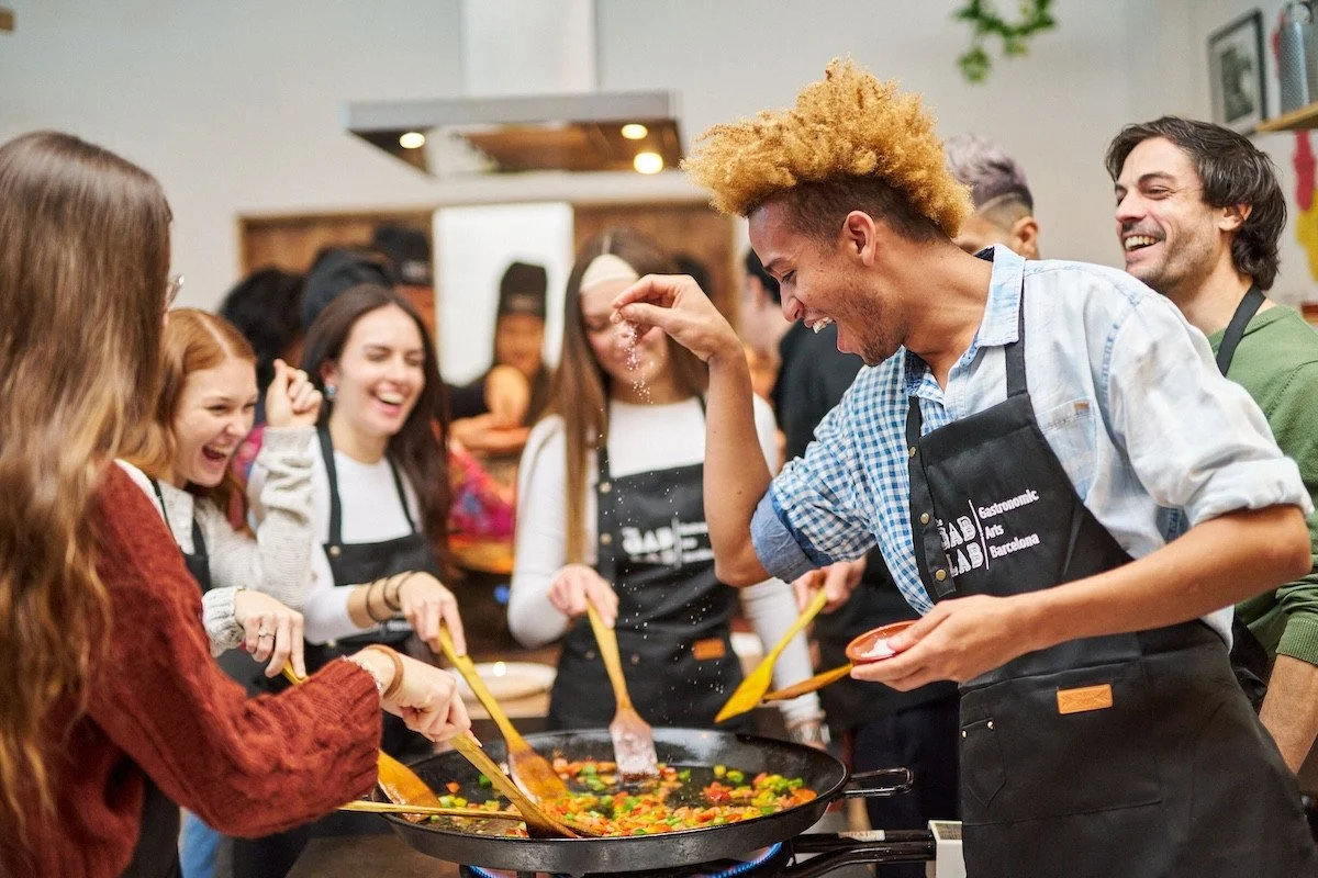 Group of people cooking together, stirring a large pan of colorful vegetables, and smiling at each other during a cooking class.