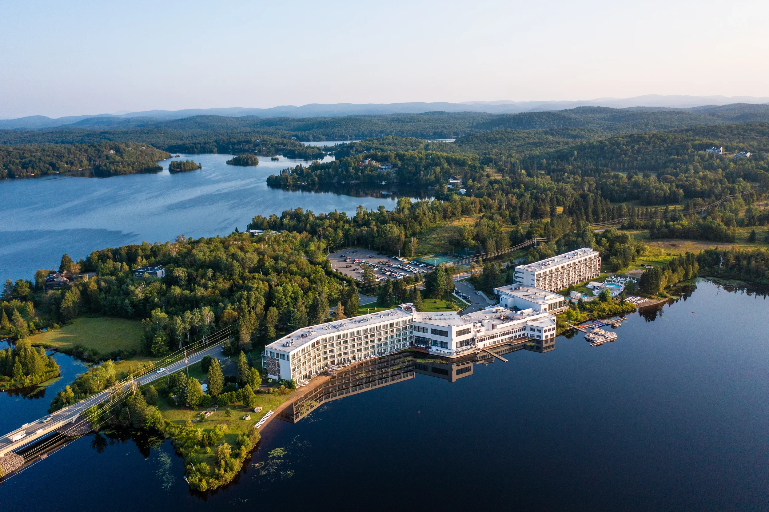 Aerial view of a lakeside resort with multiple buildings, a large parking lot, surrounded by lush green trees, with a large lake in the background and distant mountains.
