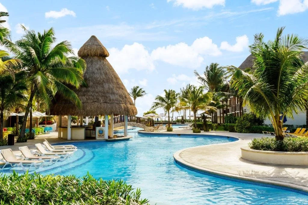 Resort pool area with palm trees, lounge chairs, thatched-roof gazebo, and a clear blue sky.