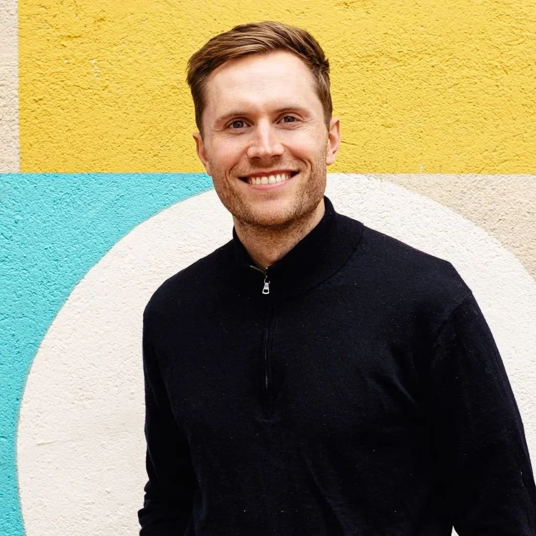 A smiling man wearing a black zip-up sweater and light blue jeans, standing in front of a colorful geometric mural with blocks of yellow, beige, blue, red, and white.