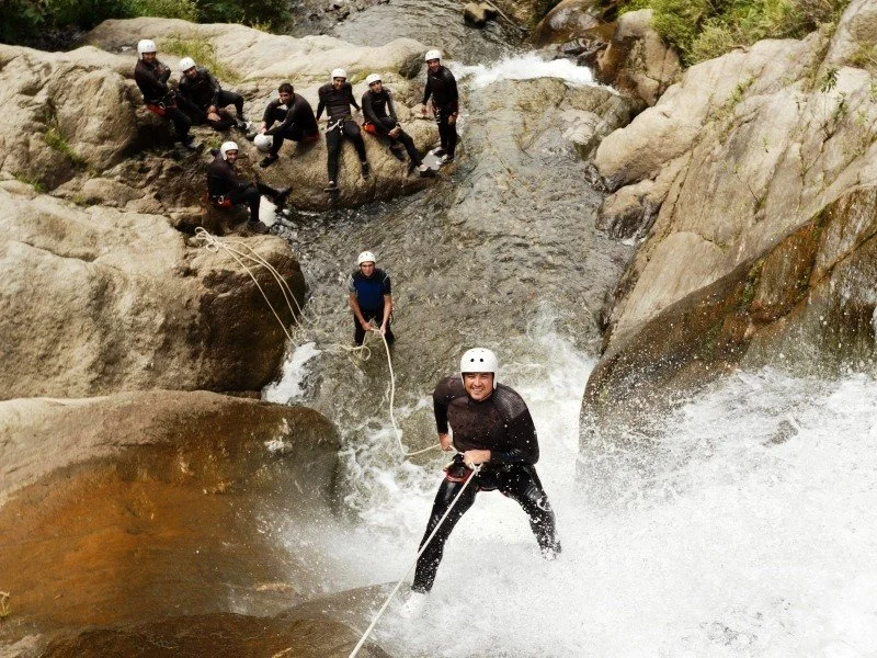 People wearing helmets and wet suits engaging in canyoning or river trekking, with some sitting on rocks and one person in the water holding onto a rope and smiling.