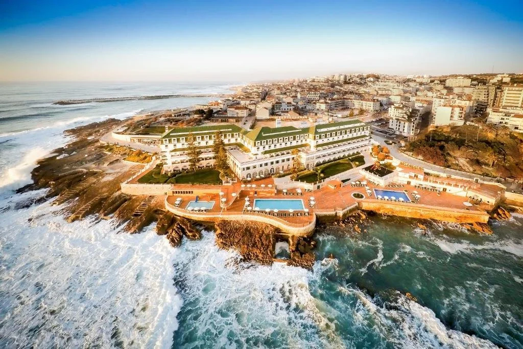 Aerial view of a large coastal hotel with multiple pools, situated on rocky shoreline with ocean waves crashing below, and a cityscape in the background.
